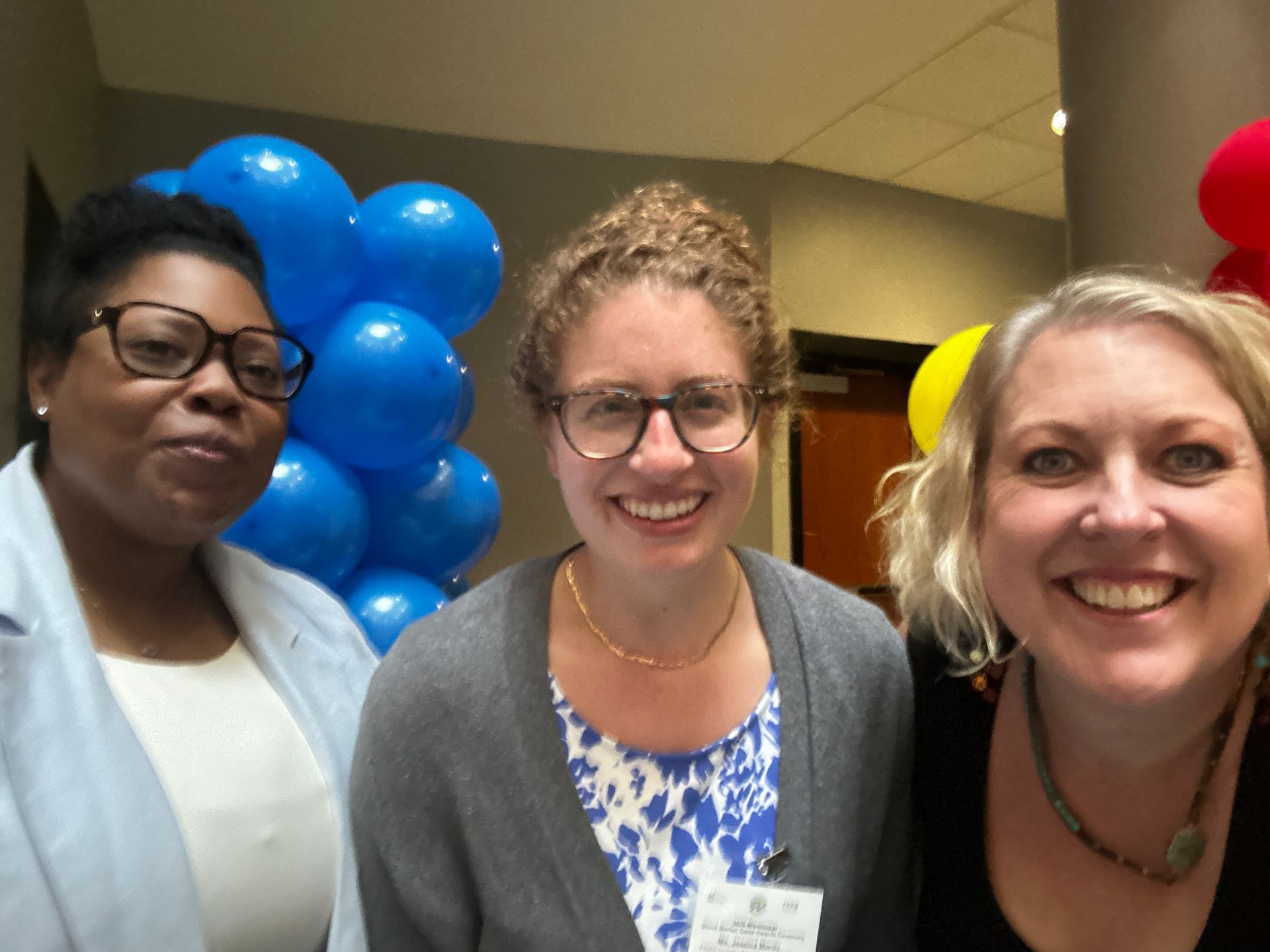 Three smiling people posing in front of blue balloons; a light-filled setting with a door in the background.