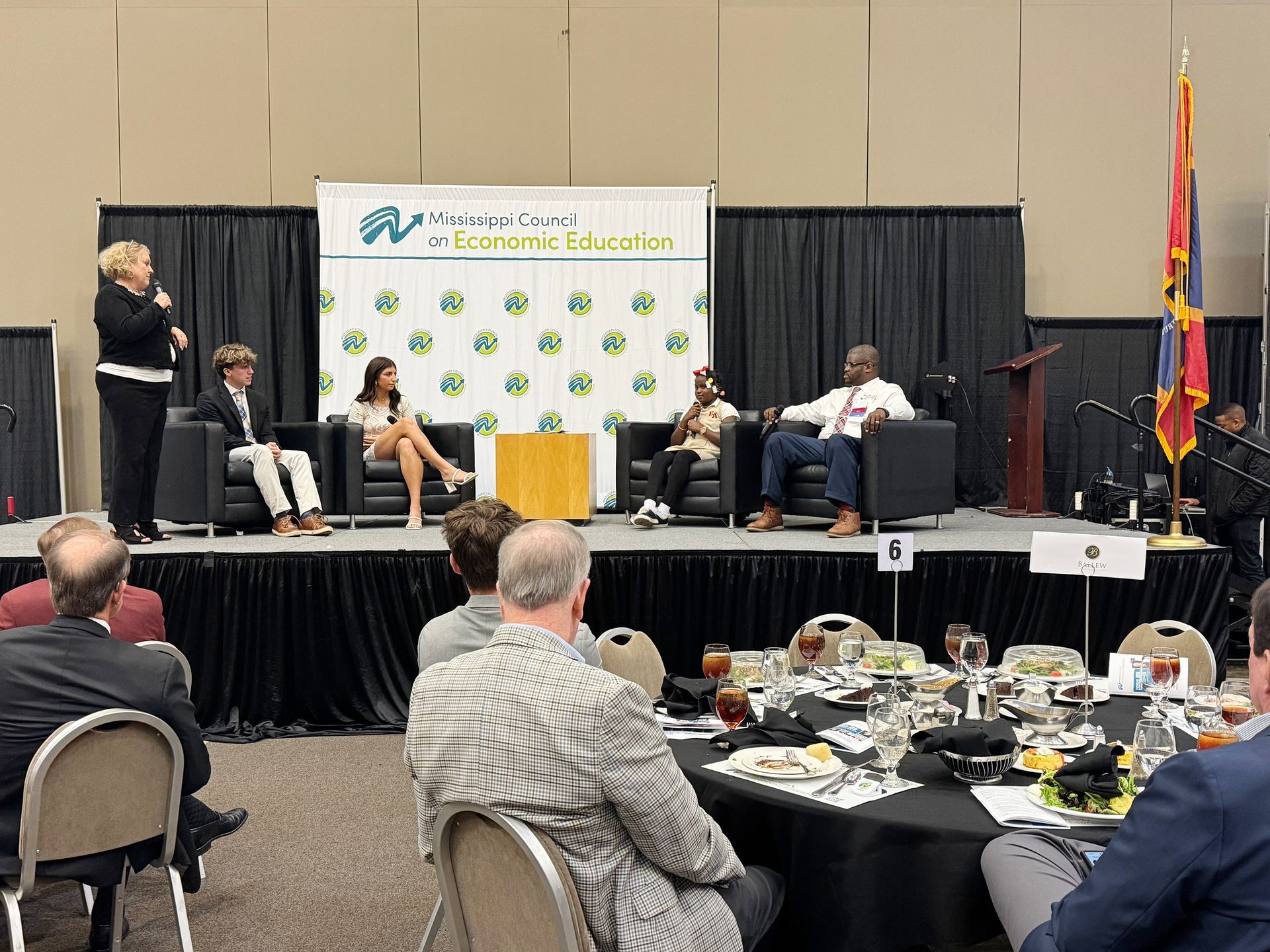 Panel discussion on stage with five people, audience in foreground. Banner with logo.