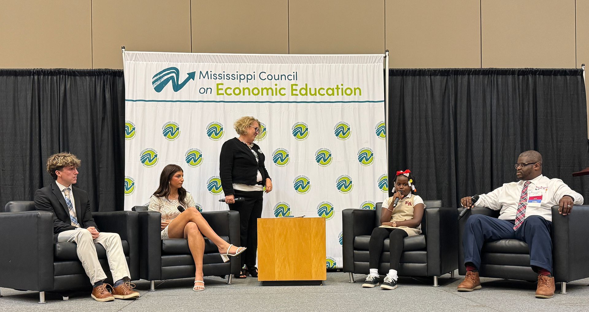 Panel discussion with five people seated, a woman standing at a podium in front of a backdrop with a logo.