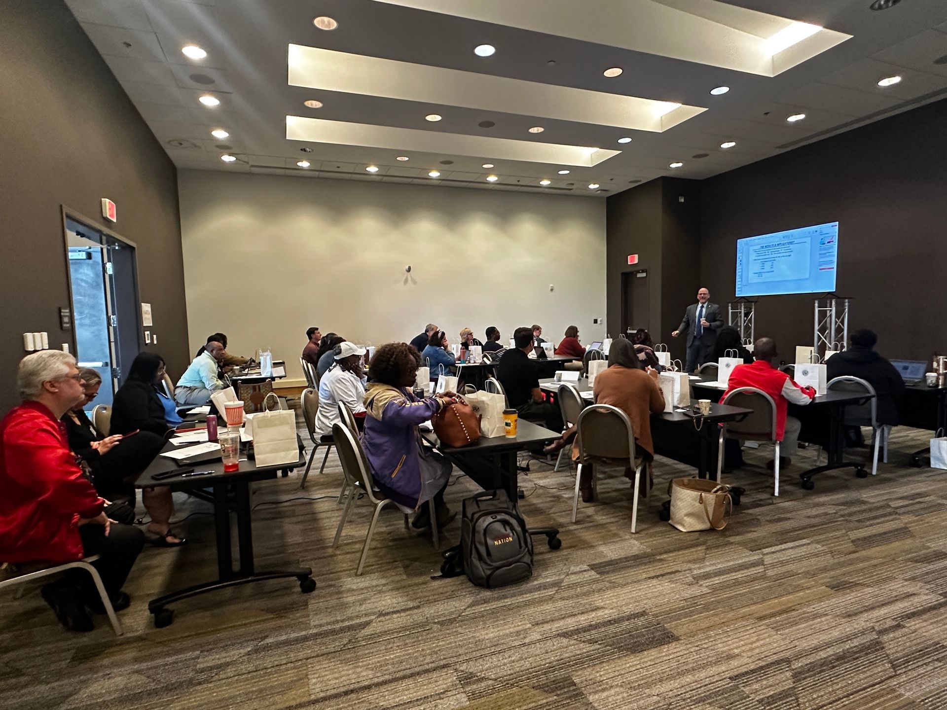 People seated at tables in a conference room listening to a presenter, with a screen in the background.