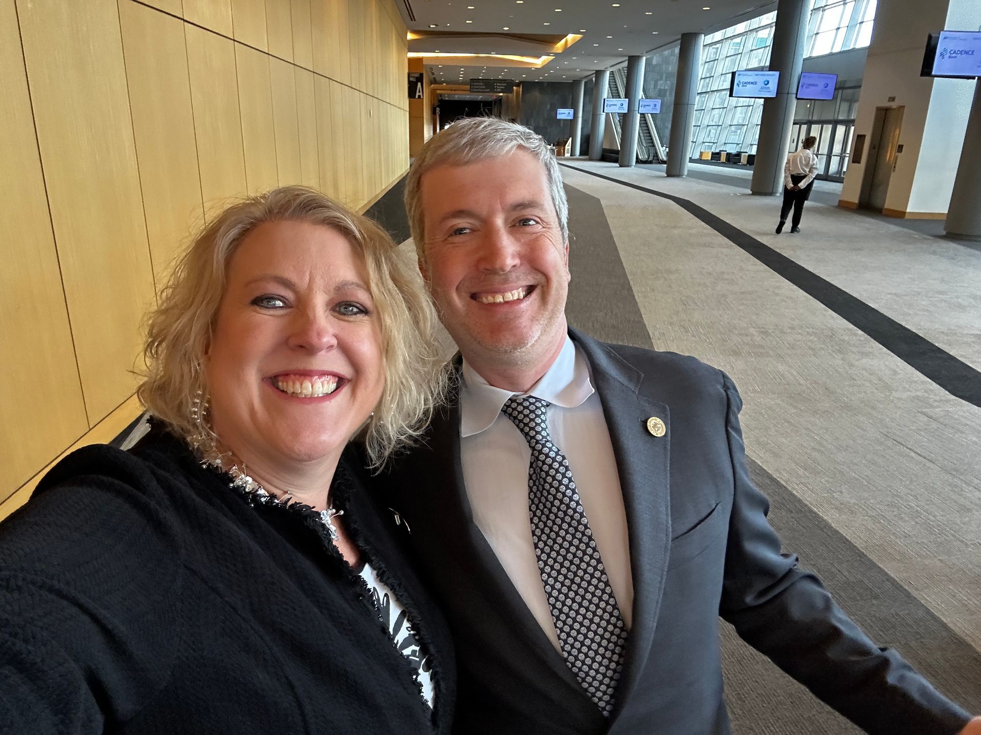 Two smiling people pose for a selfie in a modern building hallway.