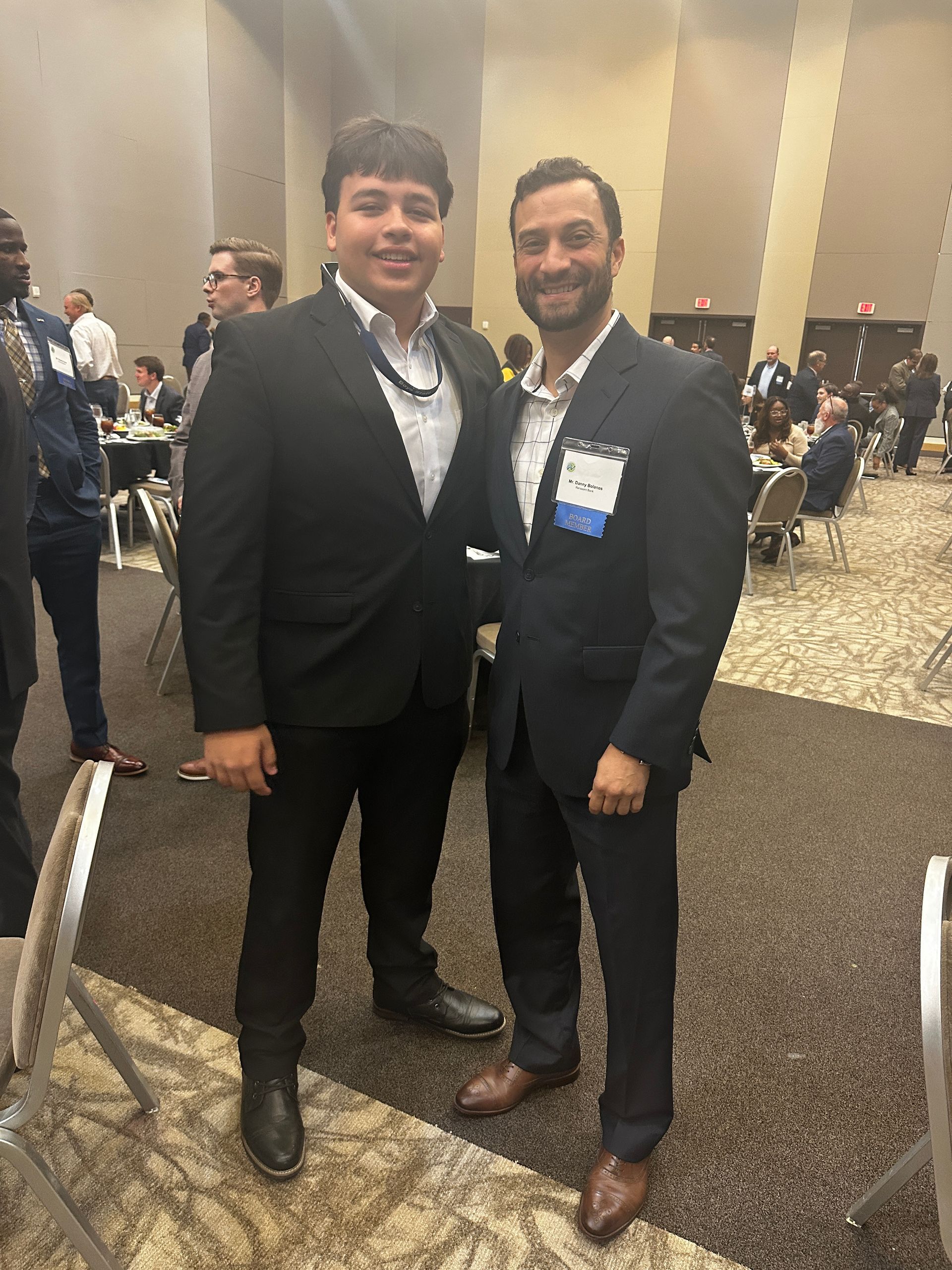 Two men in suits posing for a photo at an event, with people in the background.