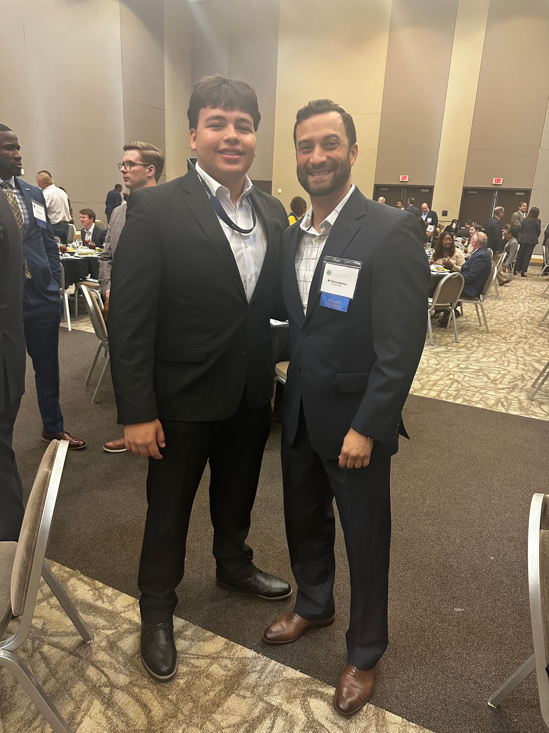 Two men in suits pose indoors at an event, smiling.