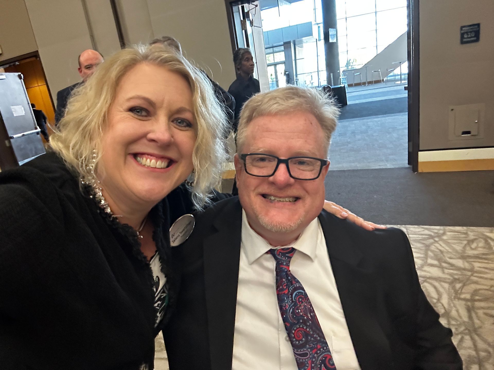 Woman and man smile for a selfie indoors. The man wears a suit with glasses. The woman has blonde hair.
