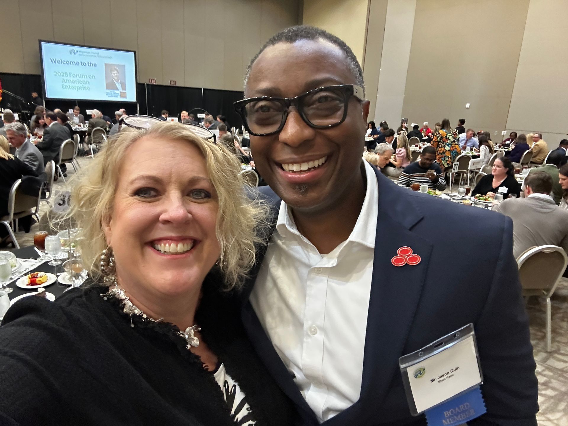 Two people smiling in a crowded room. Man in suit, glasses, woman with blond hair, posing for a selfie.