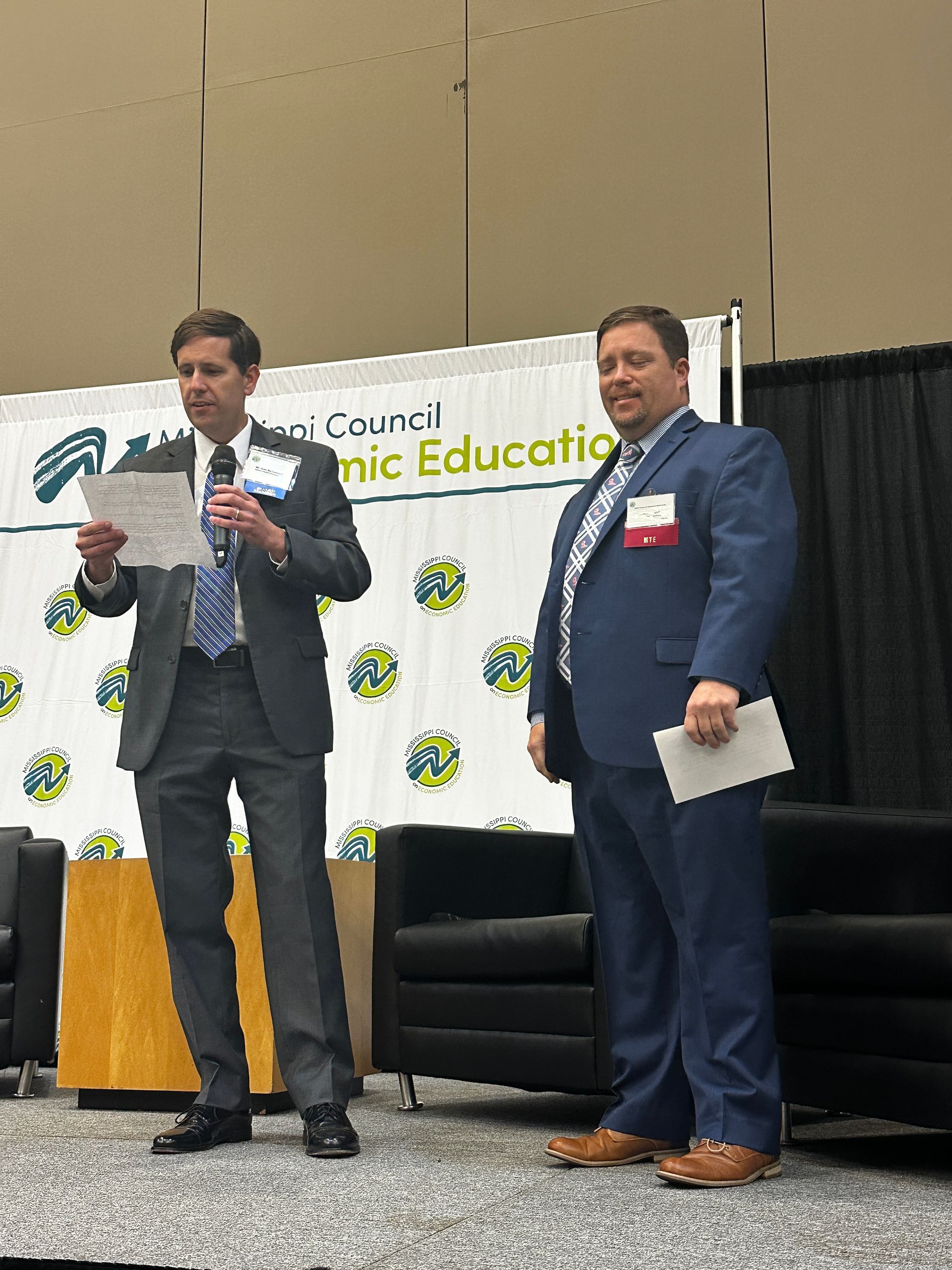 Two men in suits on a stage, one reading papers, the other holding a card. Backdrop has a logo for an education council.