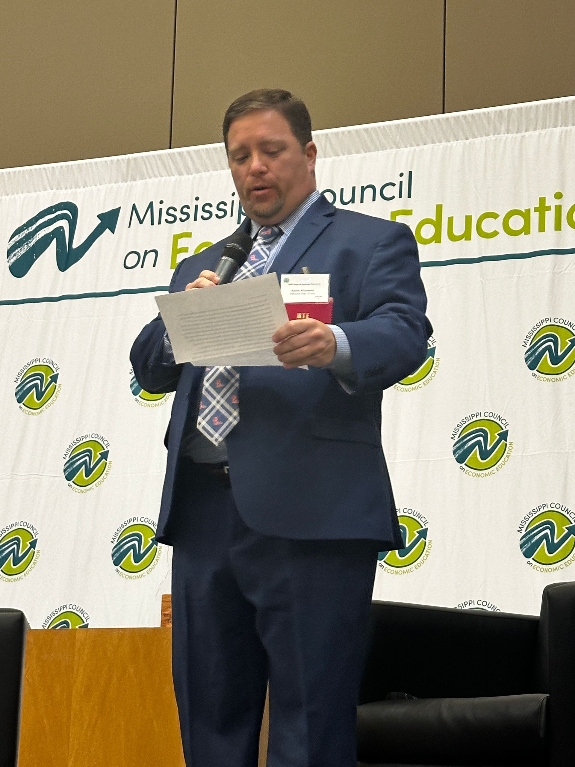 Man in a suit speaking at a conference. He is holding papers and a microphone. The backdrop has a logo.