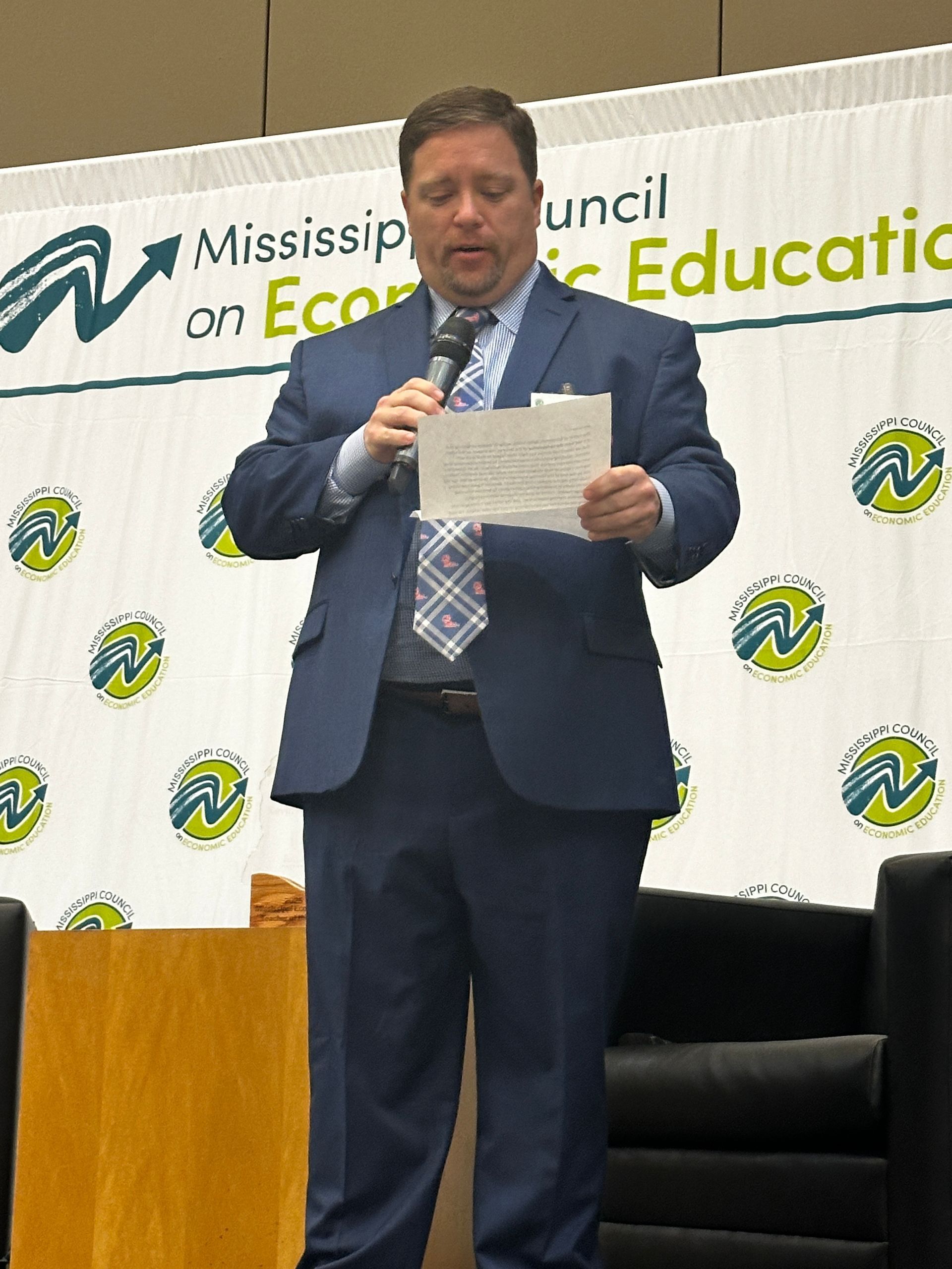 Man in suit speaking at a conference, holding paper and microphone, Mississippi Council on Economic Education banner.