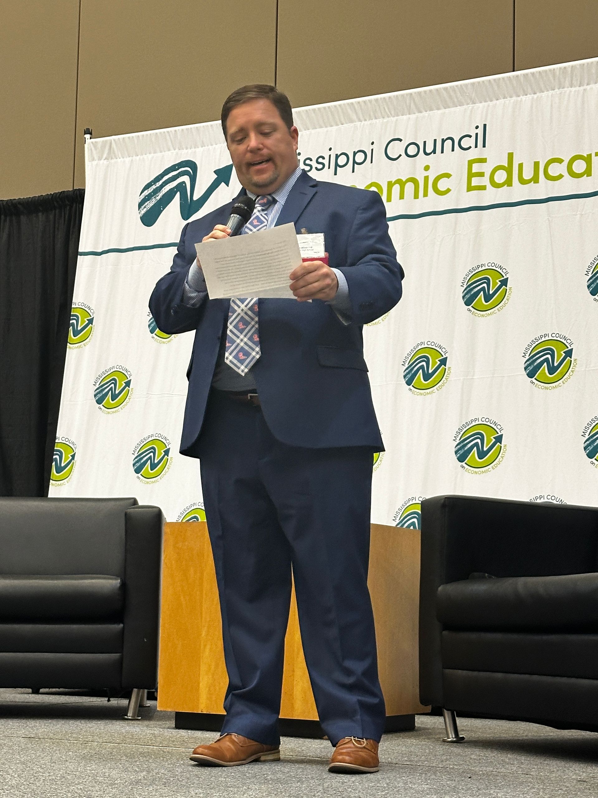 Man in a suit speaks at a podium; Mississippi Council for Academic Education banner in the background.
