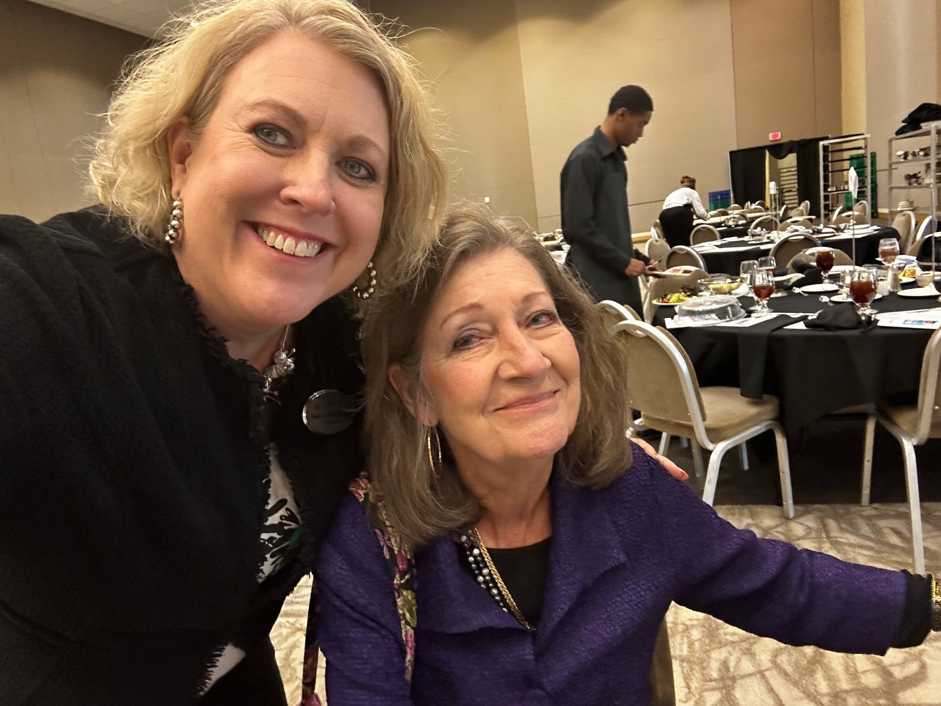 Two women smiling for a selfie at a banquet. One in black, one in purple, surrounded by tables.