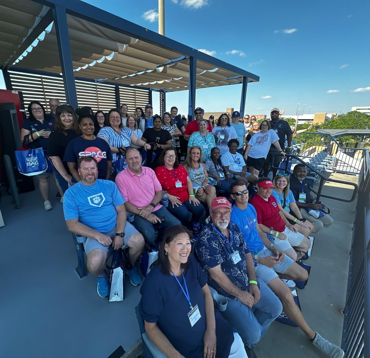 Group of people sitting and standing at a baseball stadium, under a blue roof, sunny day.