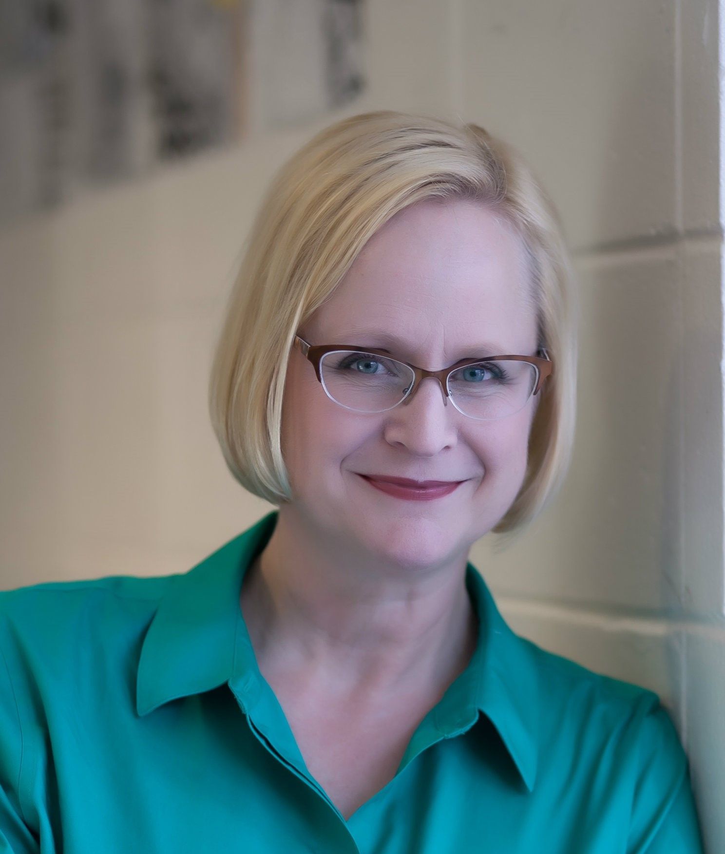 Woman with blonde hair and glasses, wearing a teal top, smiles. Leaning against a beige wall.