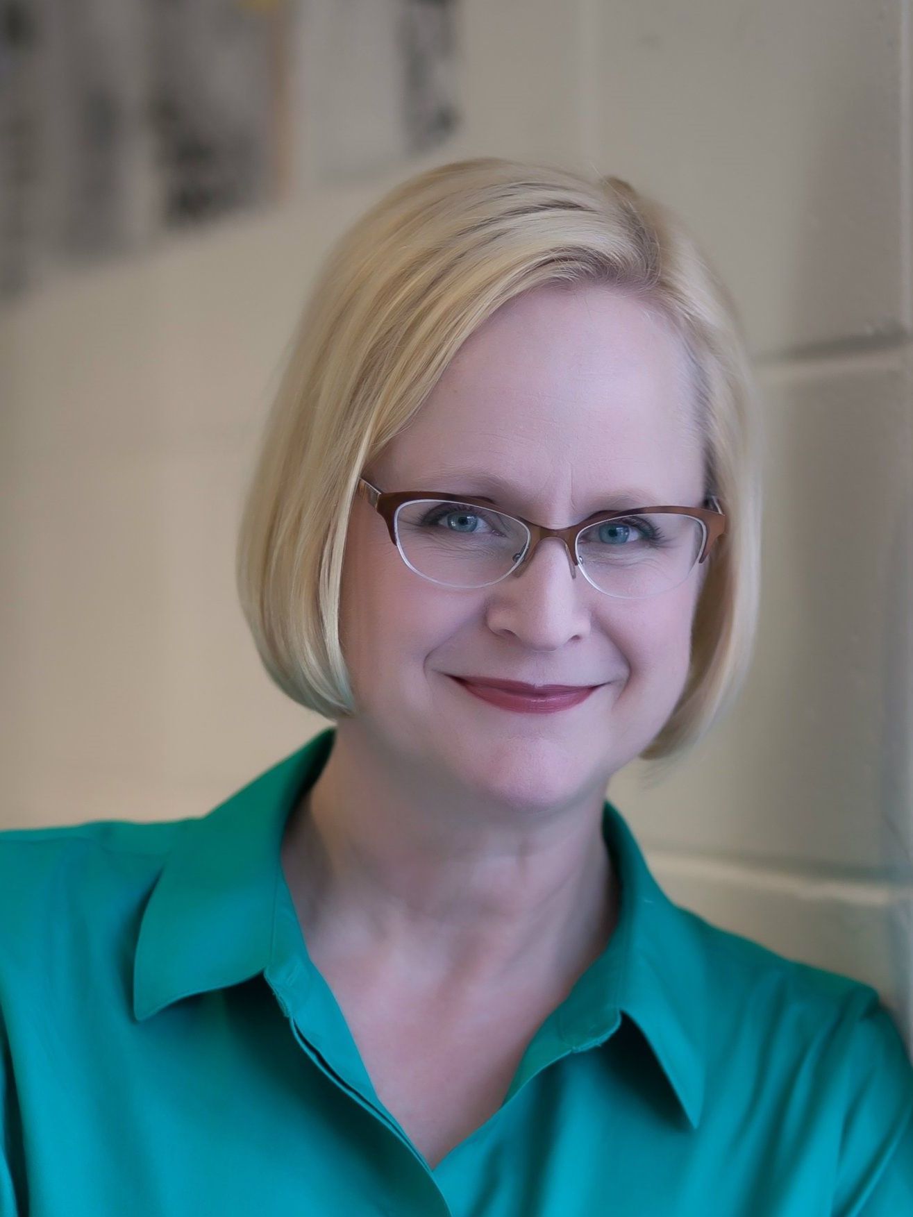 Woman with blonde hair and glasses wearing a teal top, smiling, indoors.