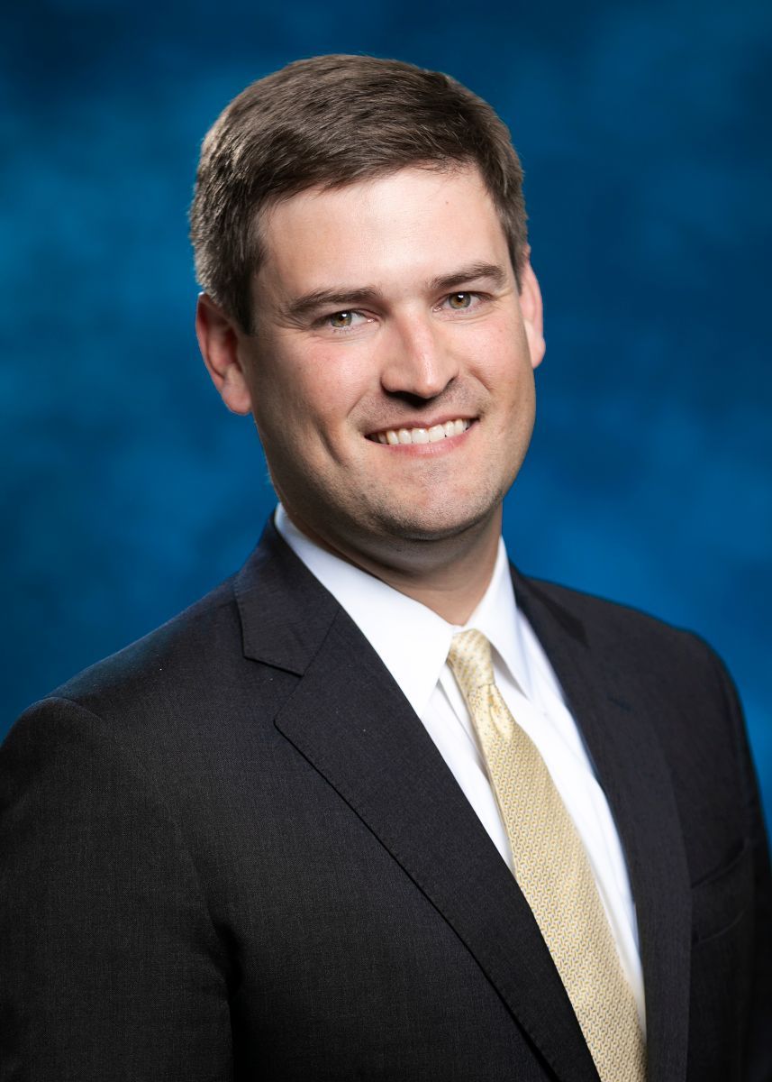 Smiling man in suit, gold tie, against a blue gradient background.