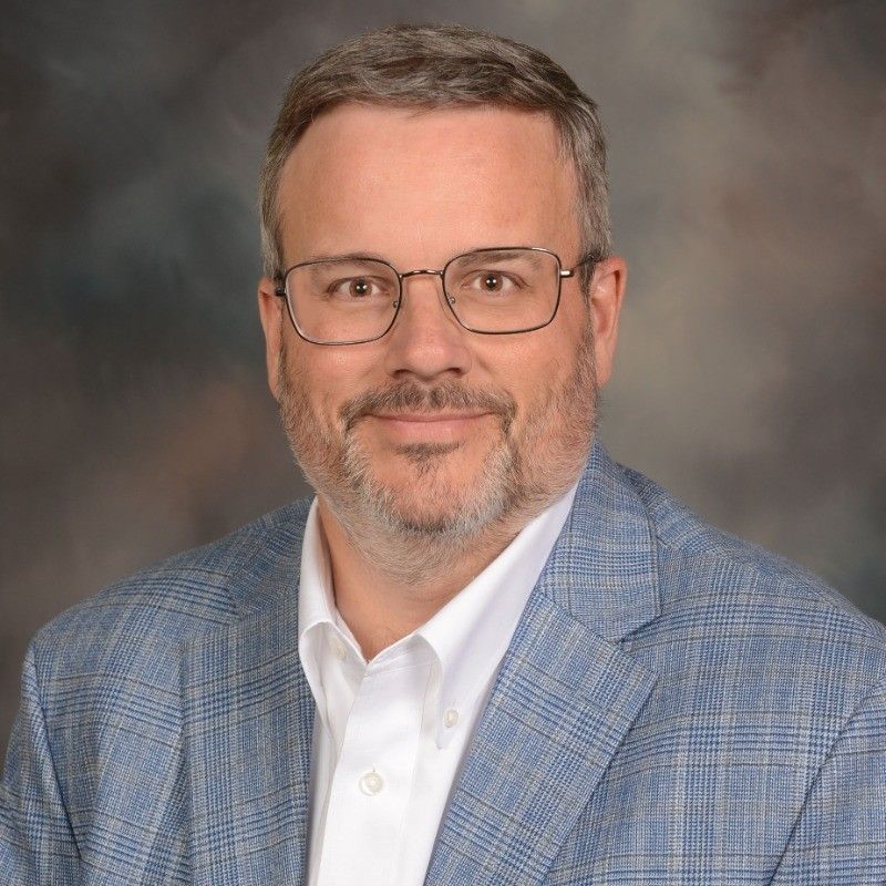 Man in a suit smiles, head and shoulders, against a blue backdrop.