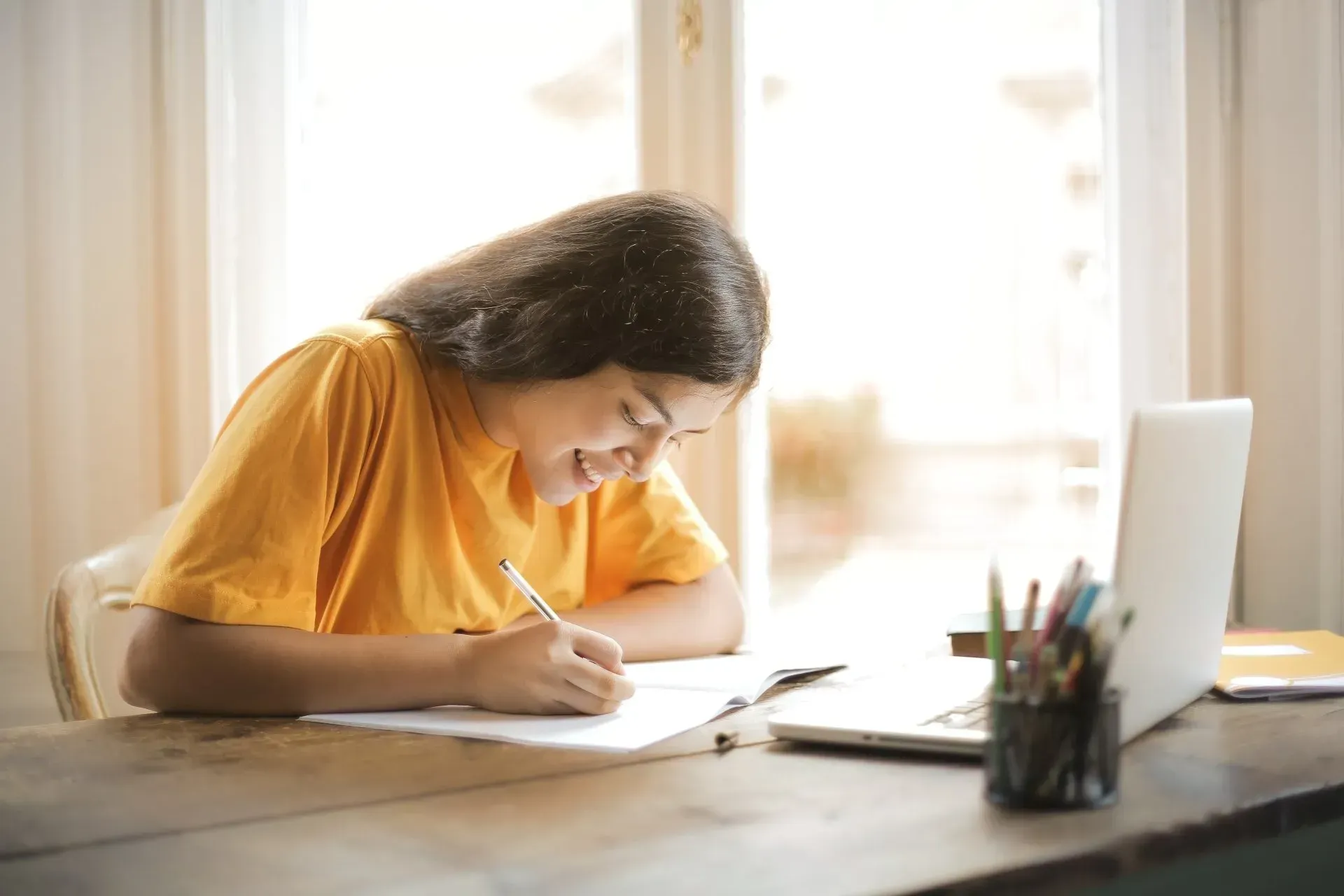 Woman smiling, writing on paper at a desk, with a laptop and pens in a bright room.