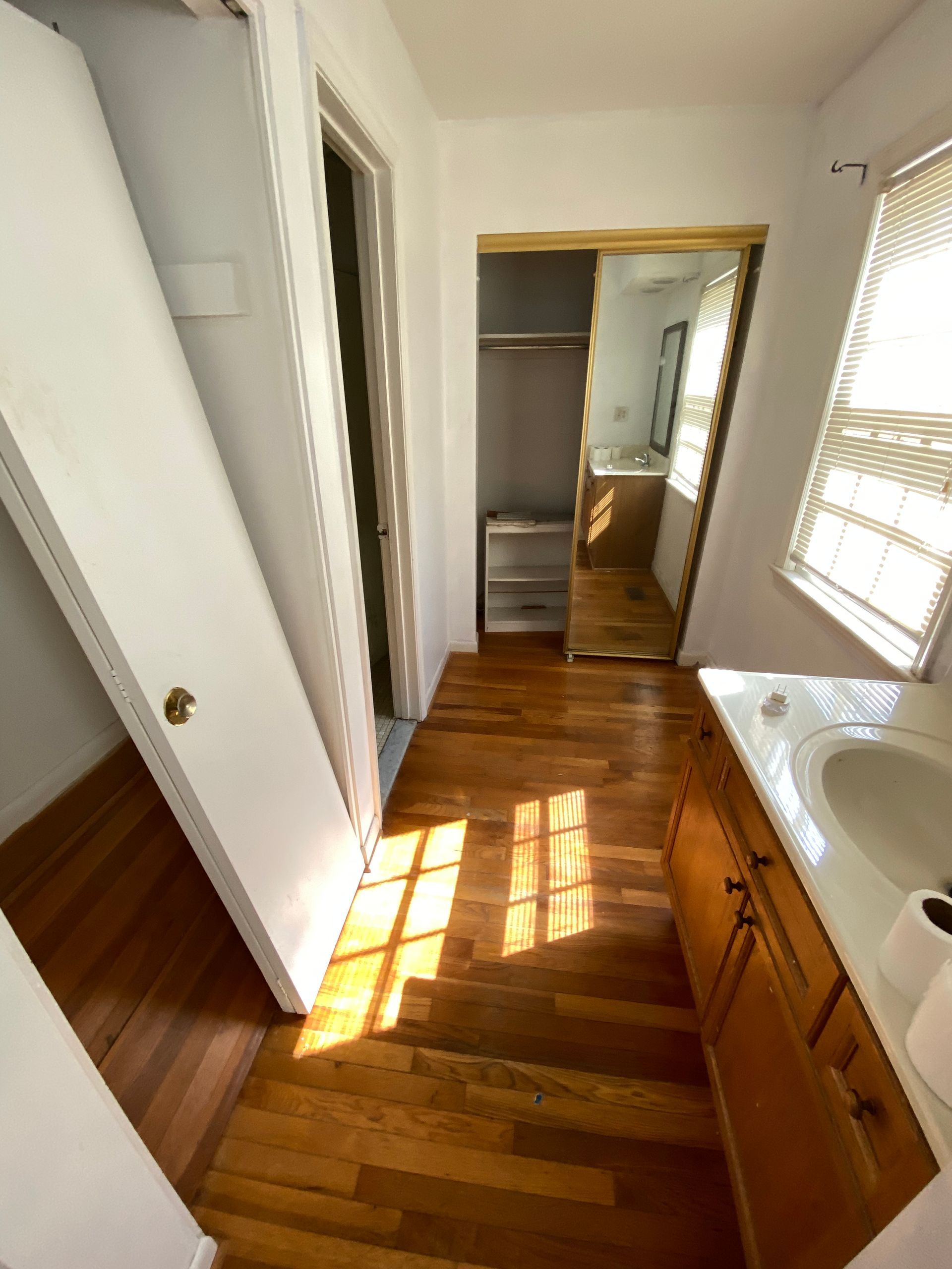 Narrow hallway with wood floor, white walls, open closet door, and sunlight streaming in from a window.
