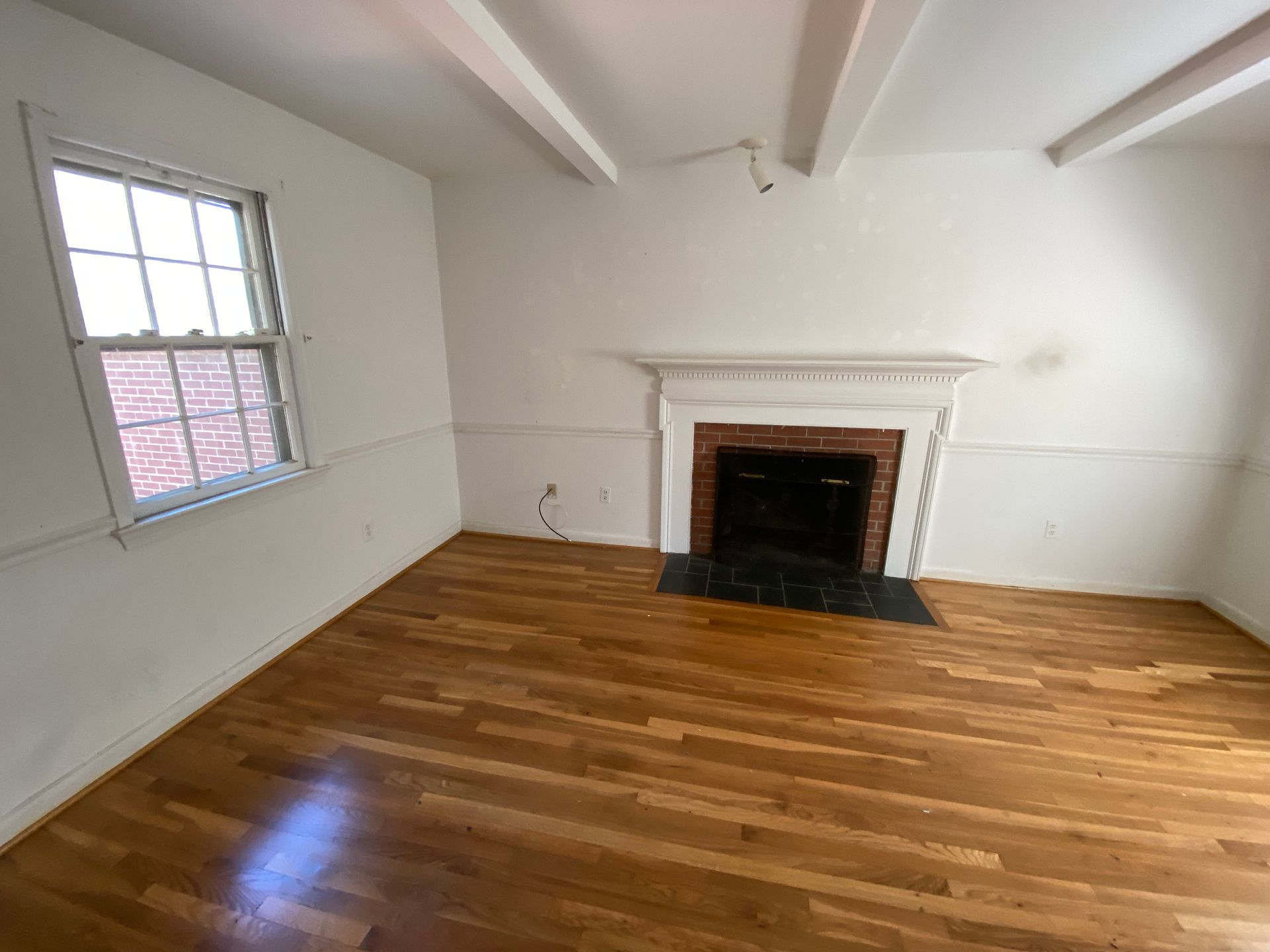 Empty room with hardwood floors, white walls, exposed beams, a window, and a brick fireplace.