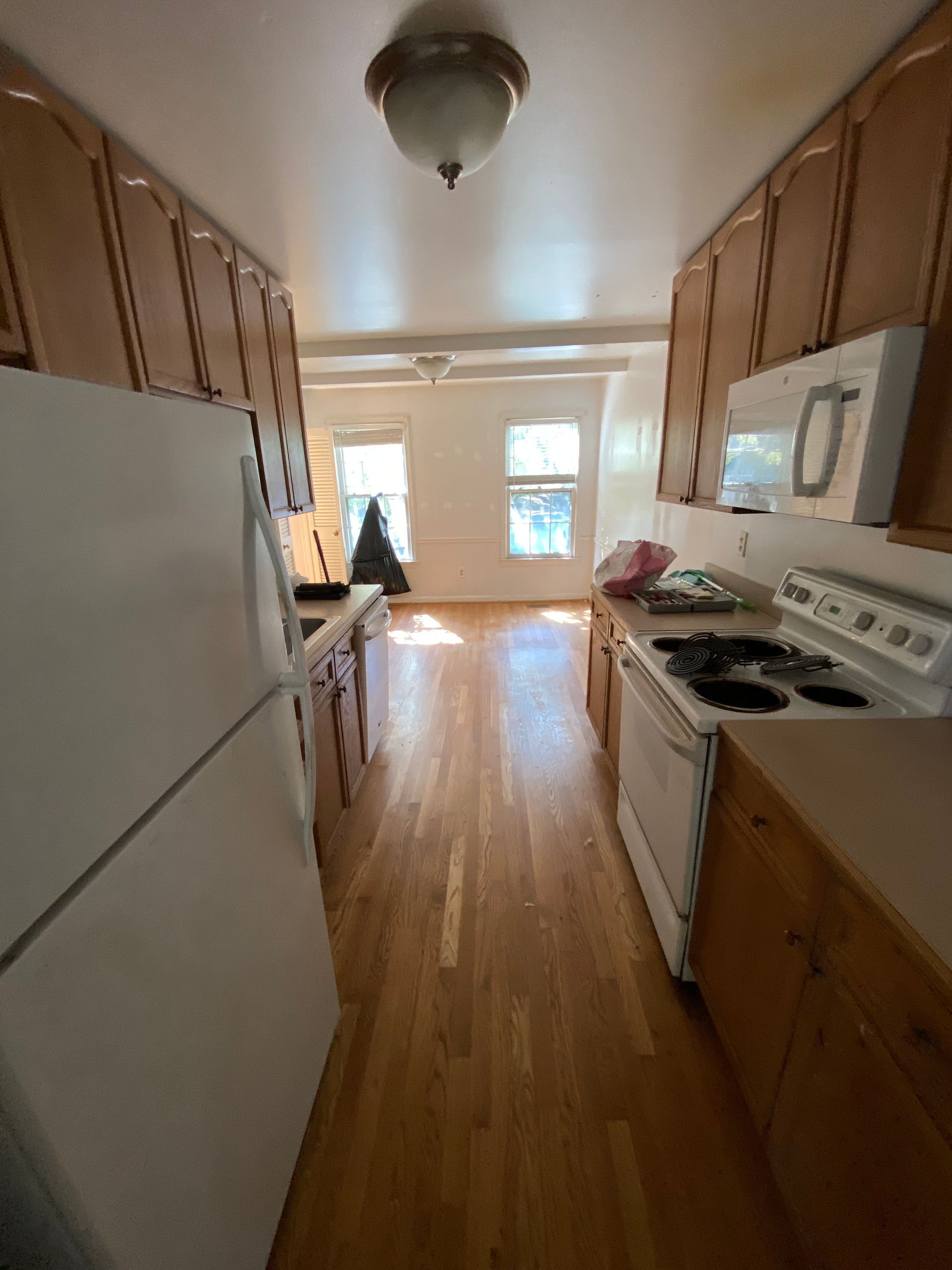 Narrow kitchen with white appliances, wood cabinets, and sunlight from a back window.