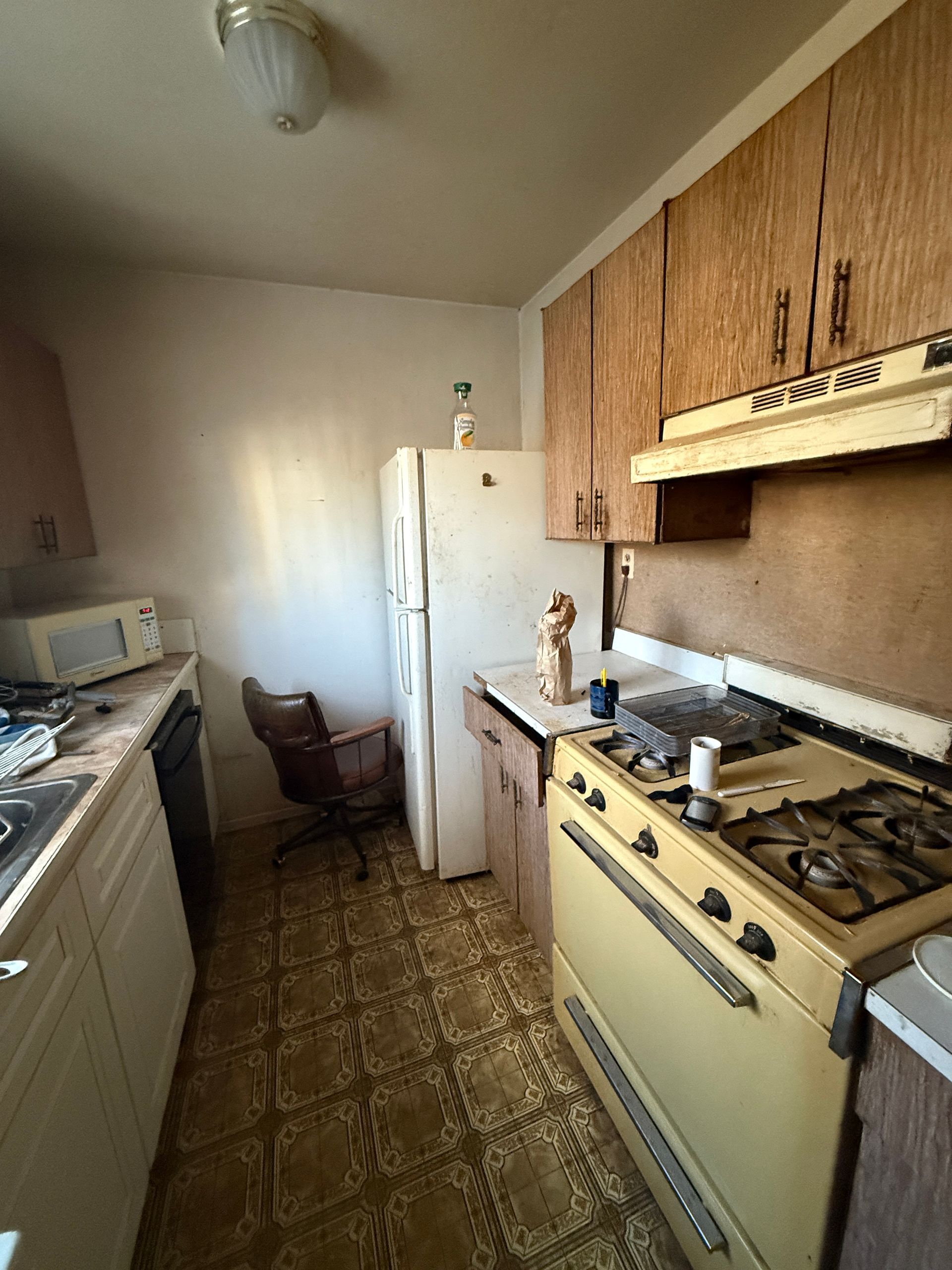 Narrow vintage kitchen with beige cabinets, white fridge, gas stove, and patterned linoleum floor.