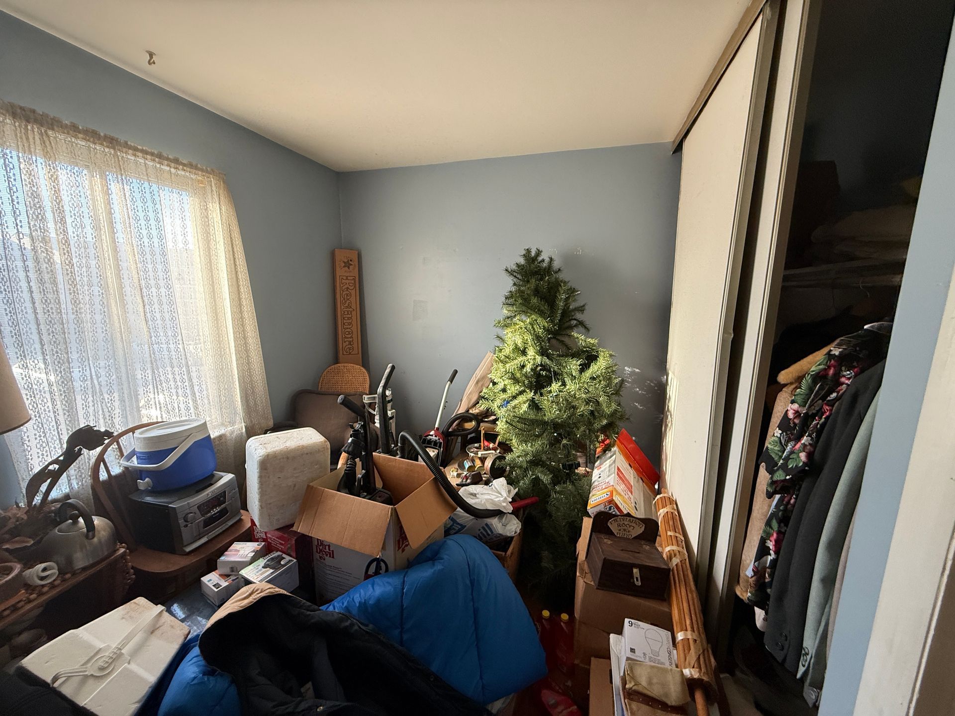 Cluttered bedroom with boxes, bags, a lit Christmas tree, and clothes by an open closet.