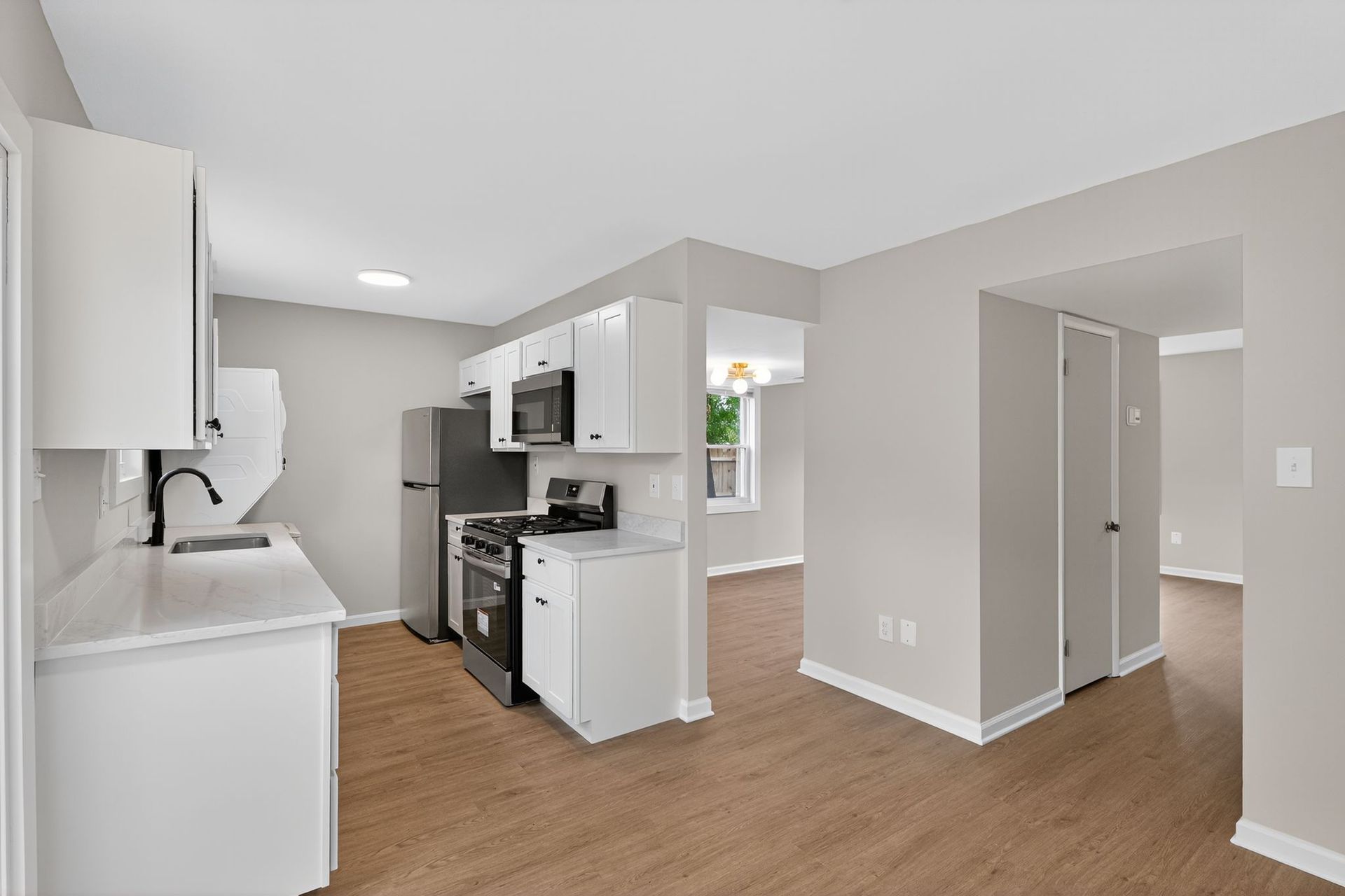 Bright empty apartment kitchen and hallway with white cabinets, stainless appliances, and wood floors