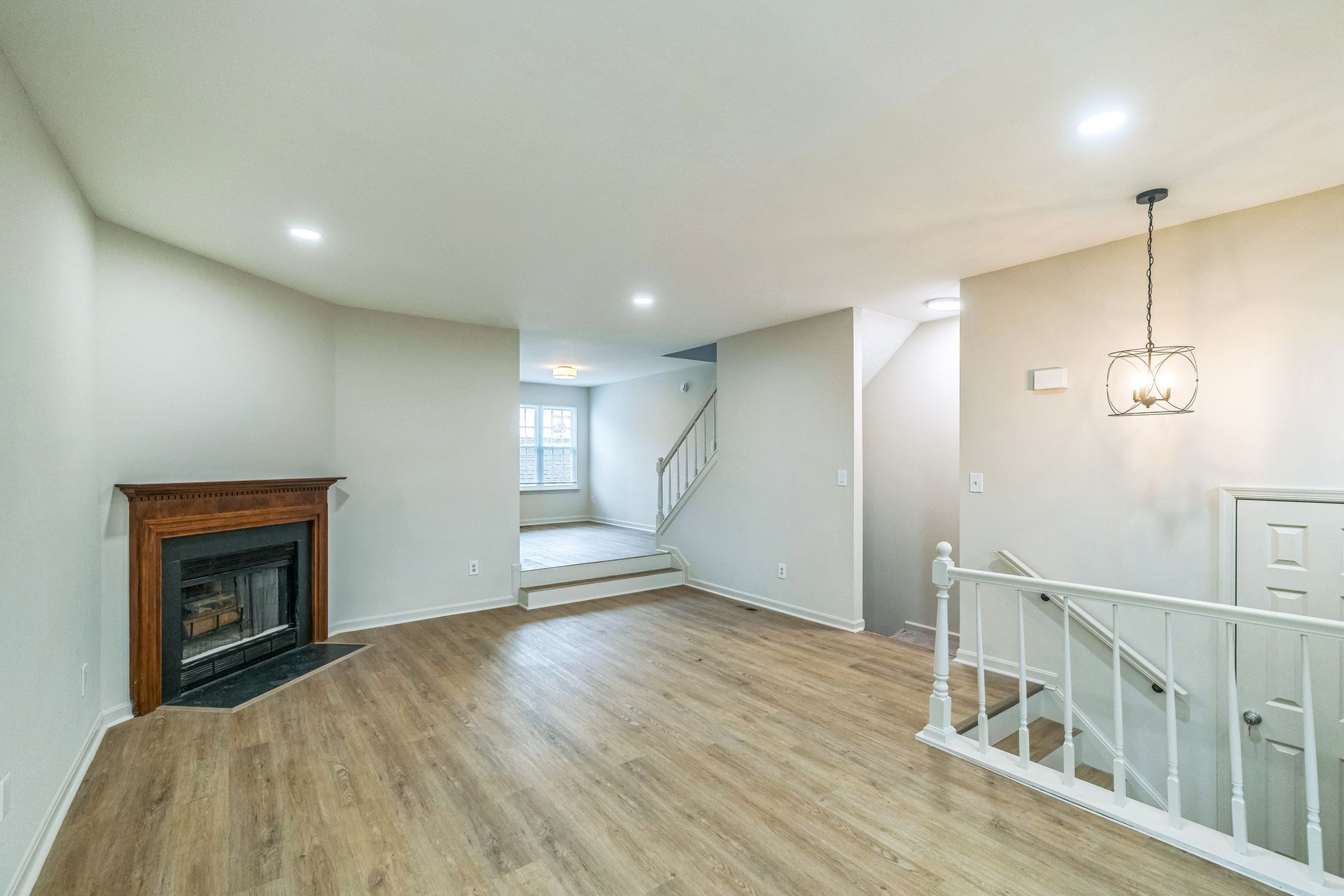 Bright open living room with hardwood floors, fireplace, white walls, and stair railing near a hallway.