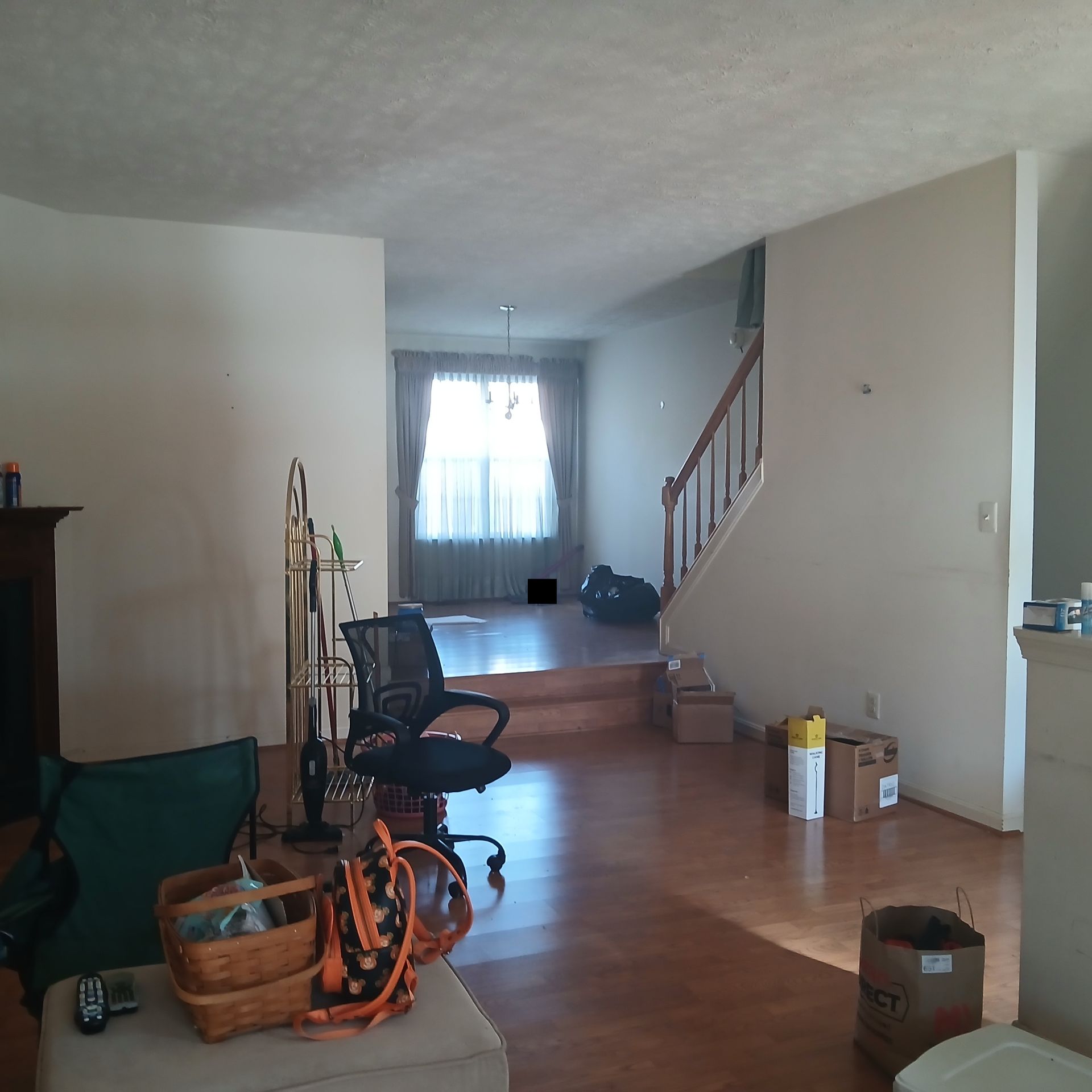 Messy living room with hardwood floor, chairs, boxes, and a staircase in the background
