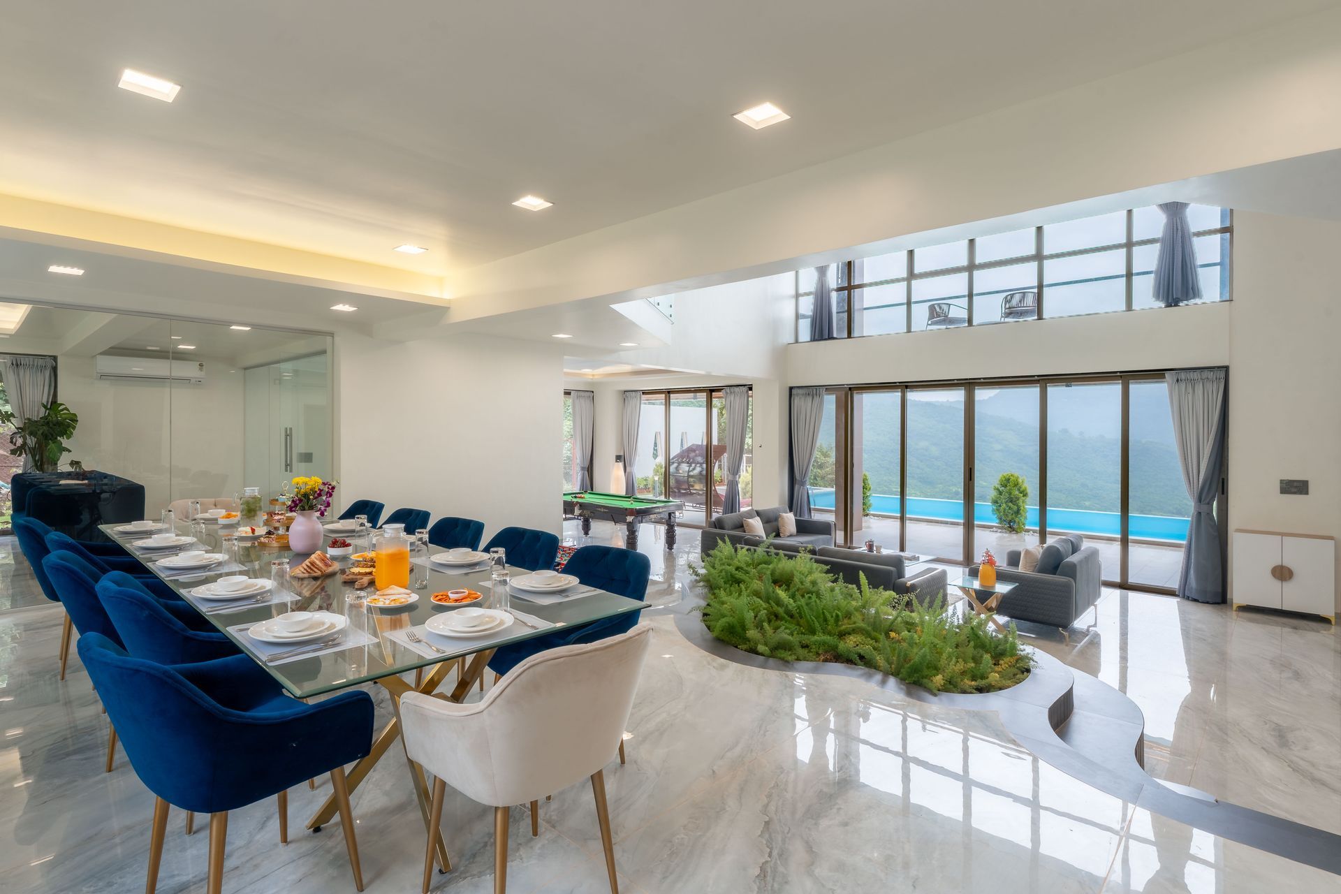 Modern dining room with blue velvet chairs, set table, indoor greenery, and large windows overlooking a bright view