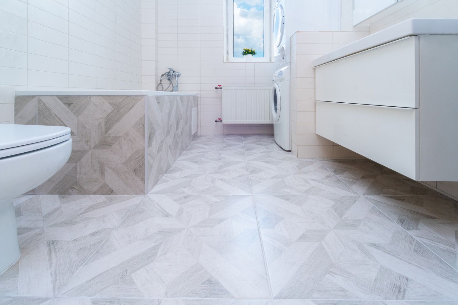 Bright modern bathroom with white tiled walls, patterned floor, bathtub, toilet, sink, and vanity cabinet