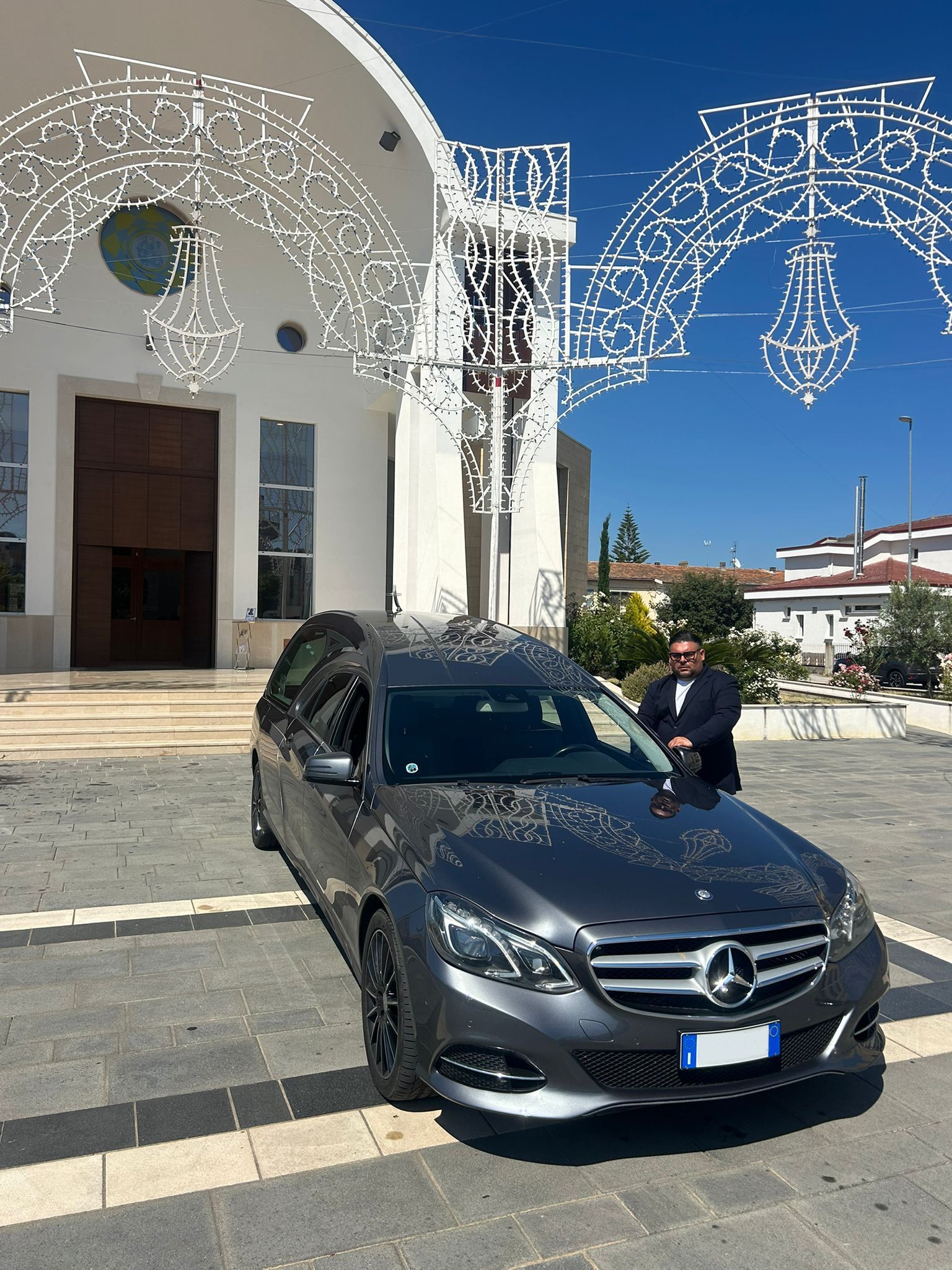 Una Mercedes grigia è parcheggiata davanti a un edificio bianco illuminato da luci decorative; un uomo è in piedi accanto ad essa, sotto un cielo azzurro.