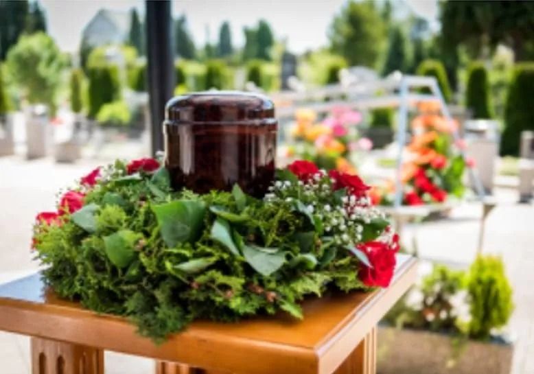 Monumento funebre cimiteriale con urna scura su una corona di fiori in cima a un supporto di legno, circondato da tombe sfocate e vegetazione.