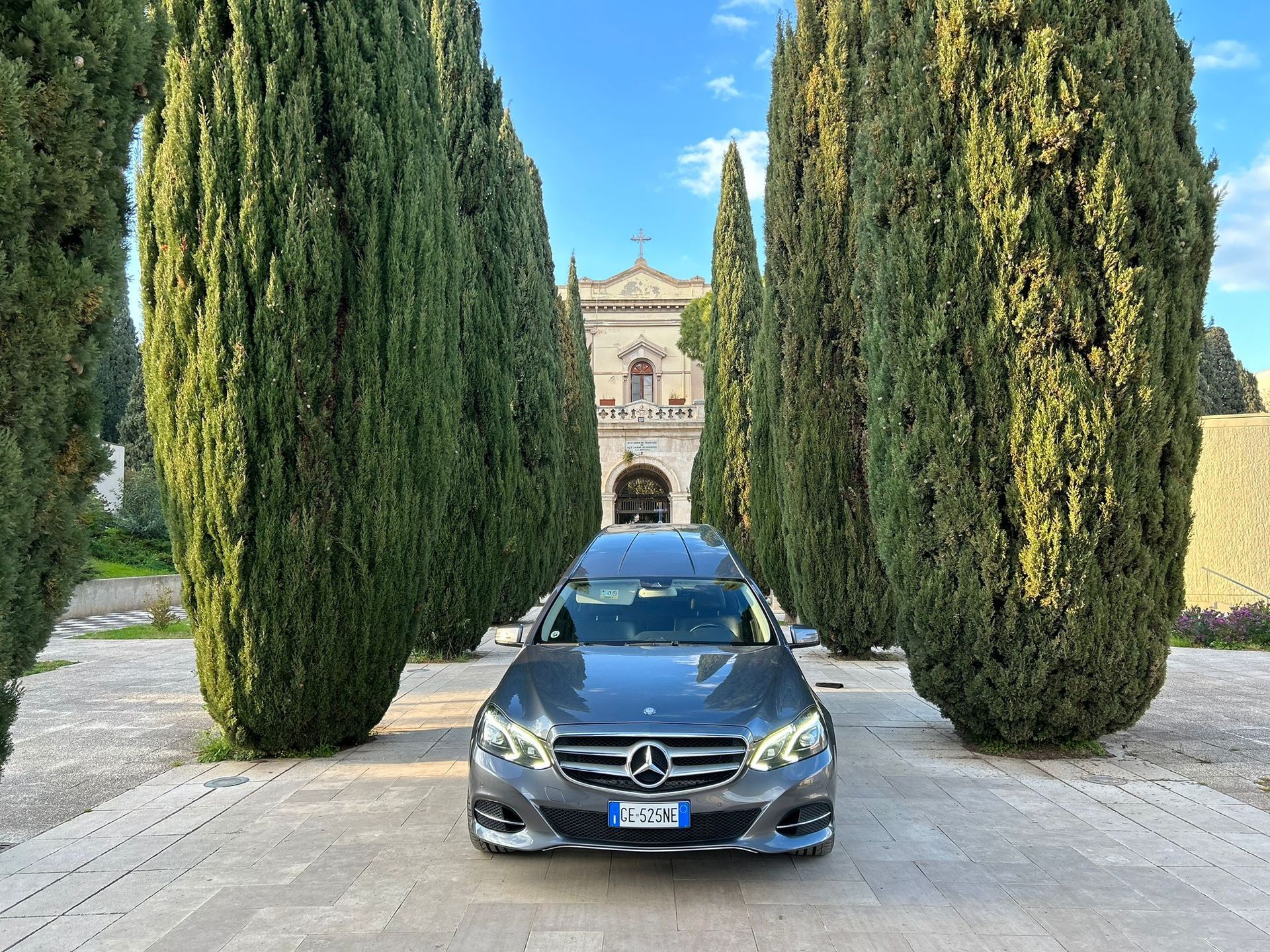 Una berlina Mercedes nera parcheggiata su un vialetto di pietra tra alti cipressi, con una villa sullo sfondo.
