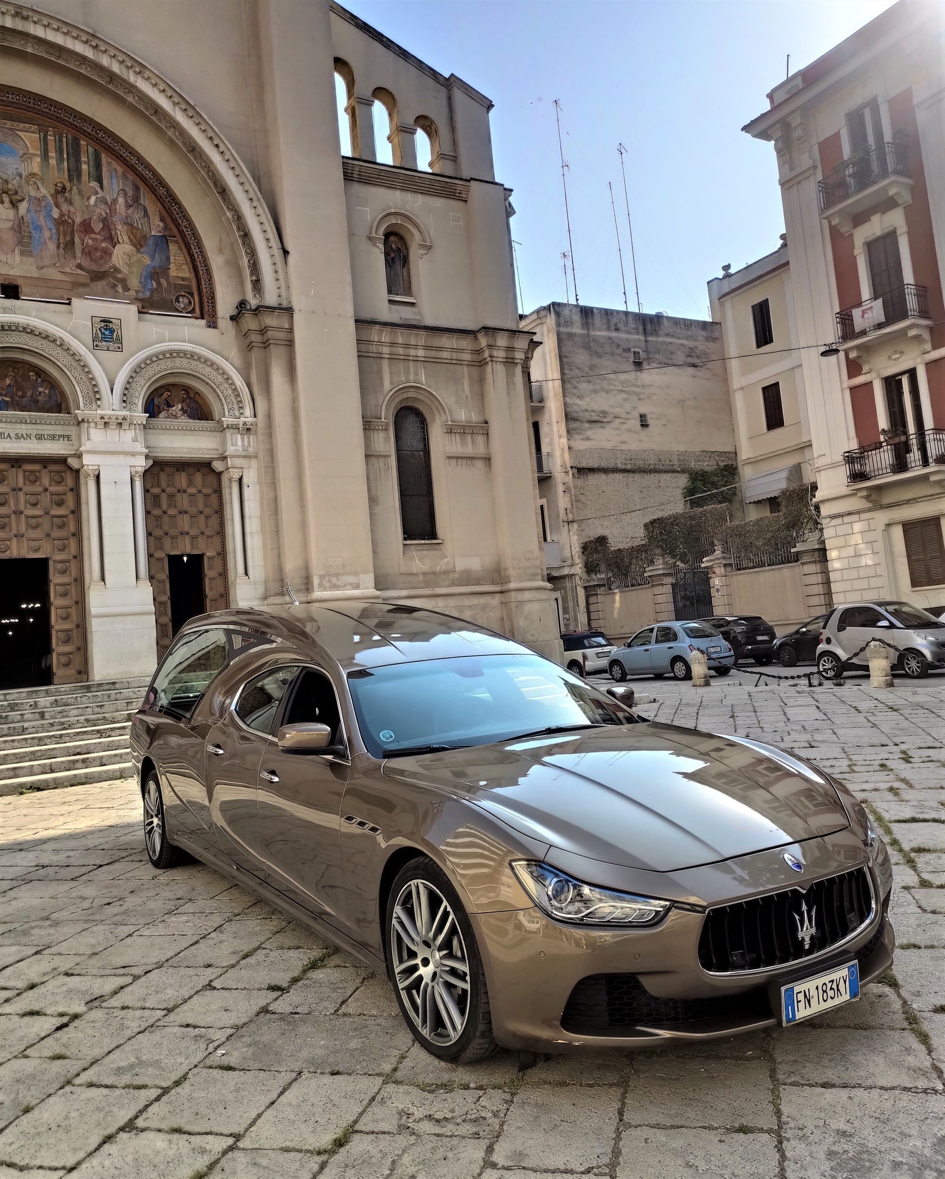 Auto di lusso marrone parcheggiata in una piazza lastricata di ciottoli accanto a edifici storici in pietra e alla facciata di una cattedrale.