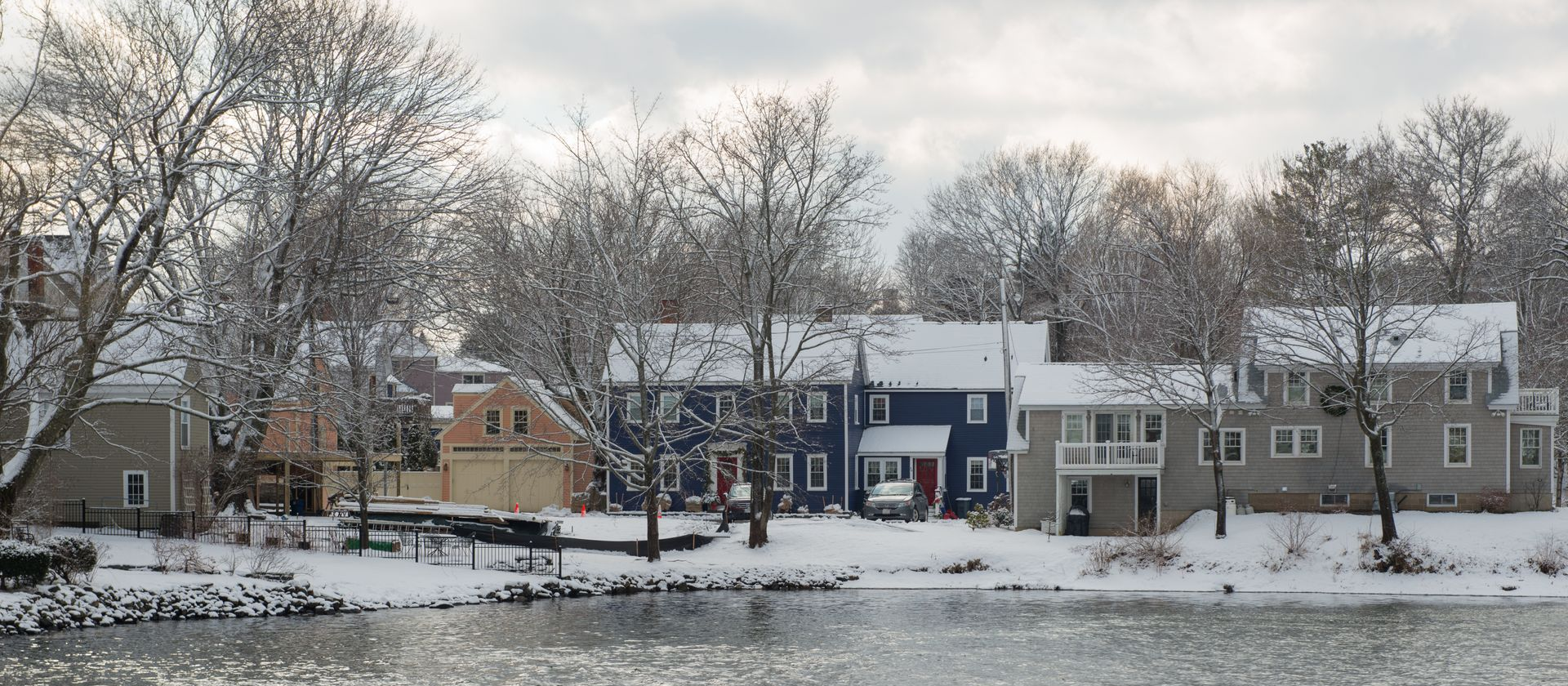 Snowy riverside neighborhood with houses and leafless trees under a winter sky