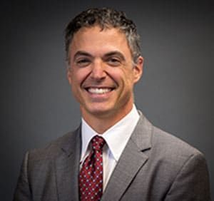 Man in gray suit and red patterned tie smiling at the camera. Dark background.