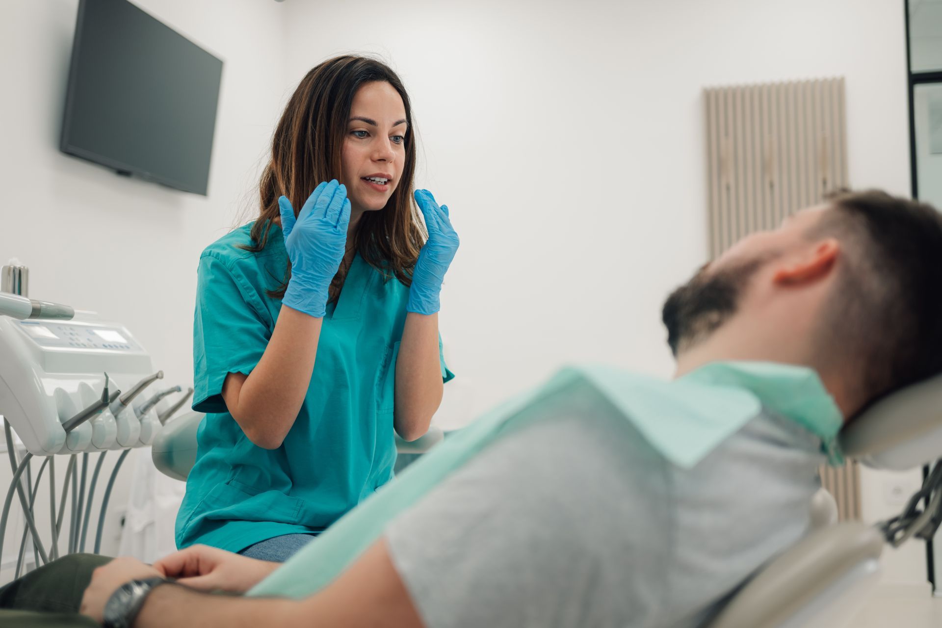 Dentist in blue gloves talks to a reclined patient in a dental exam room