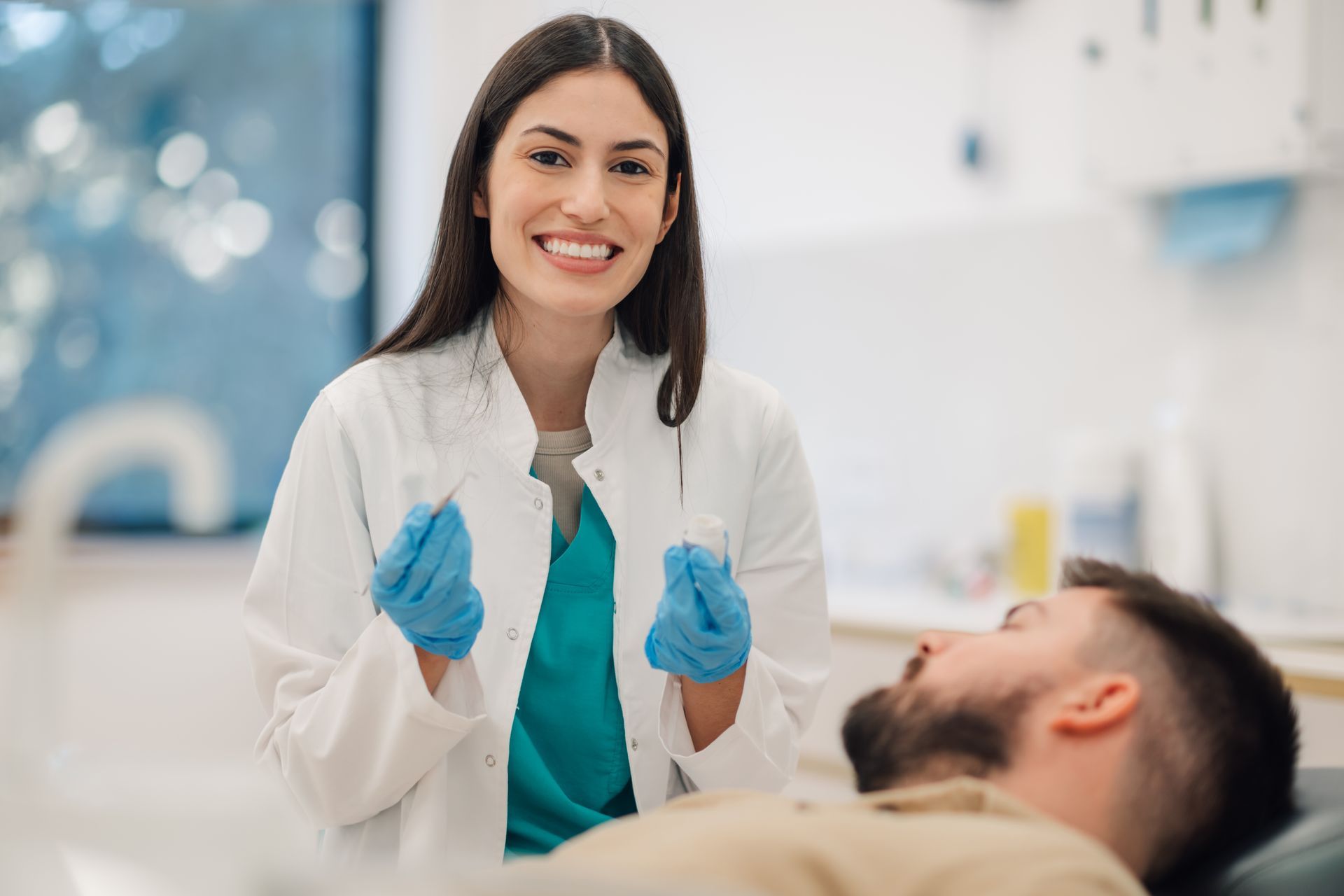 A dentist in a white coat and blue gloves smiles while holding dental tools over a patient lying in a clinical chair.