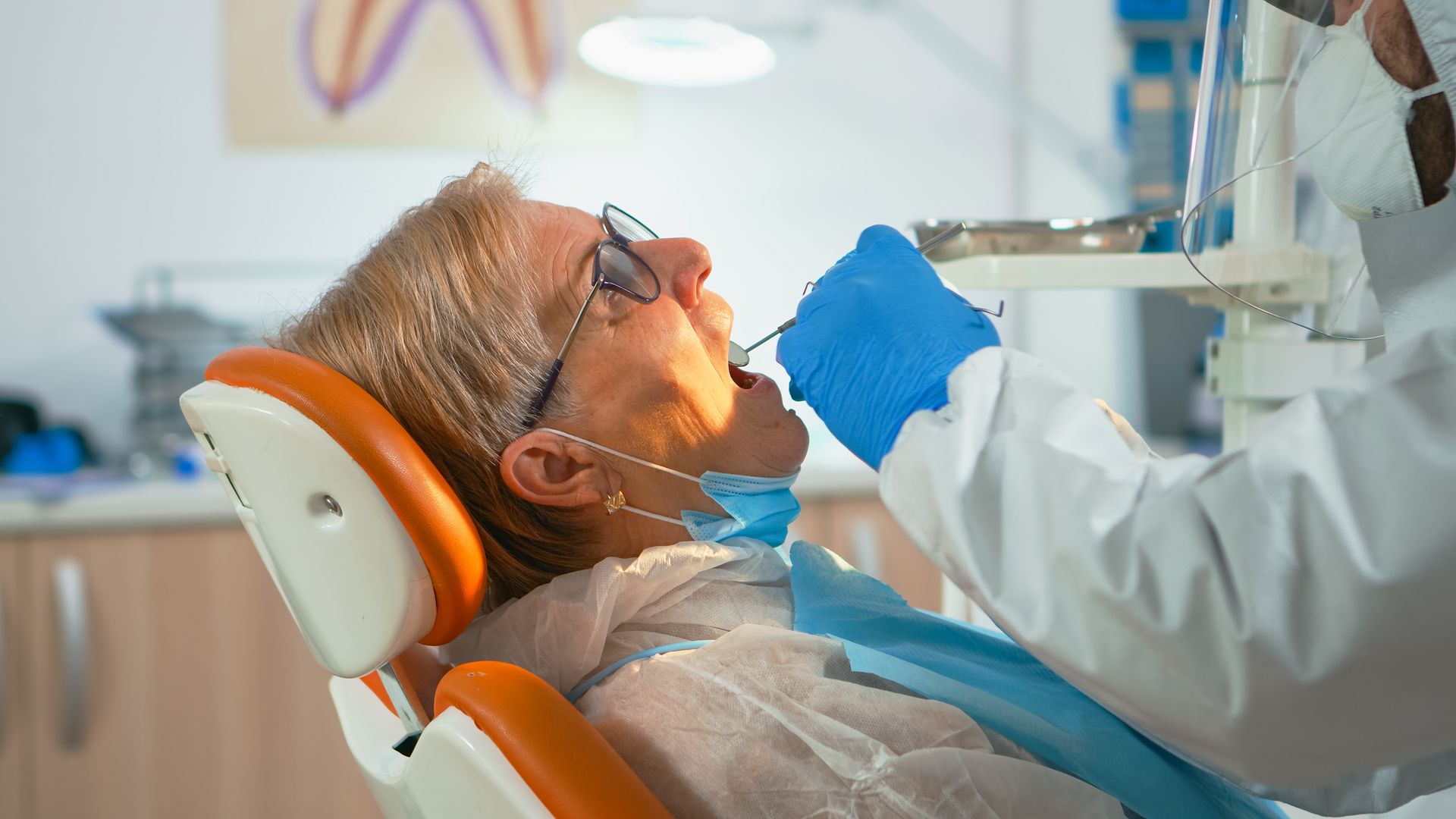 Dentist examining patient's teeth in a dental chair. Blue gloves, tools, bright lights, medical setting.