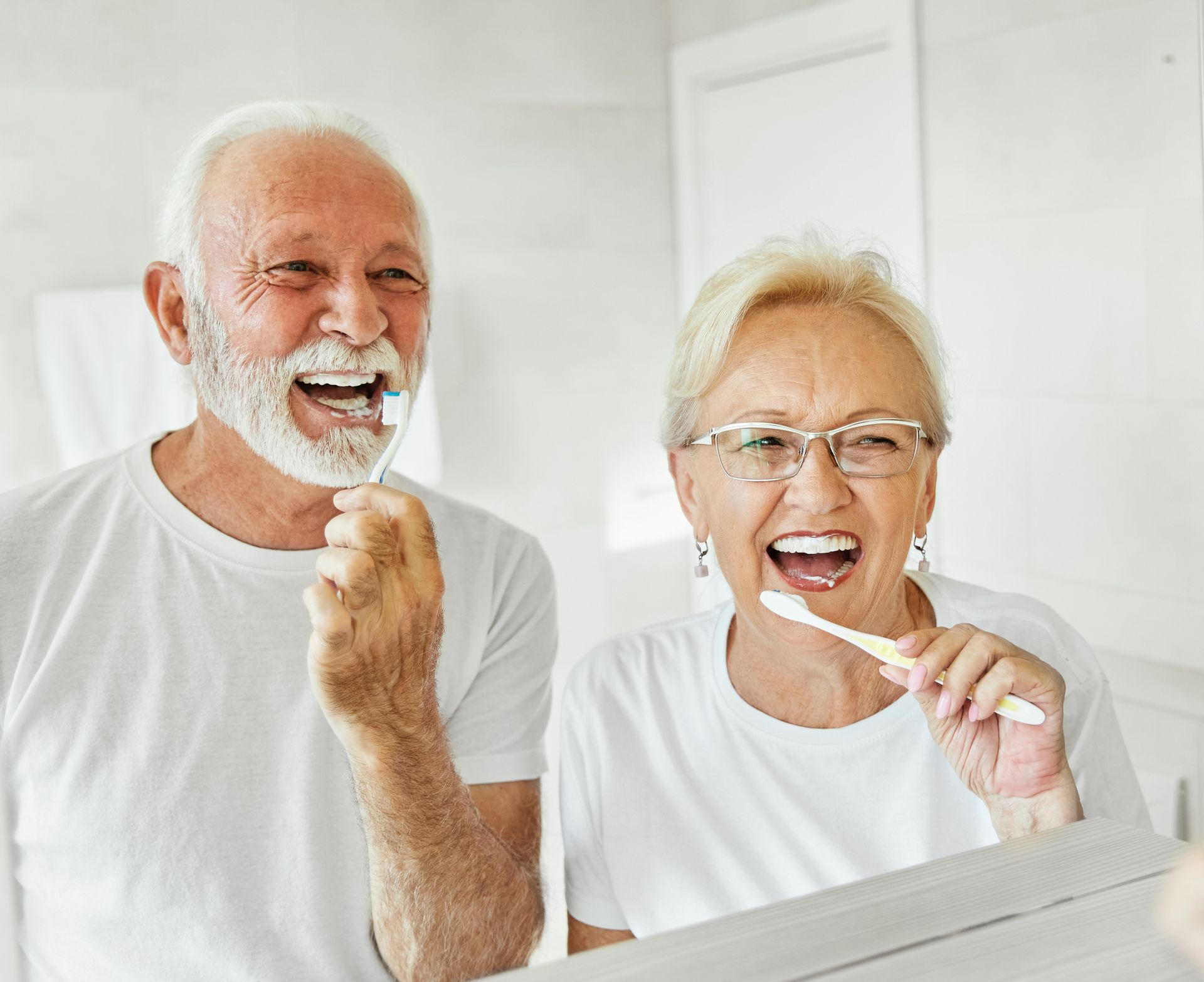 Two smiling people brushing their teeth in a bright bathroom, wearing white shirts.