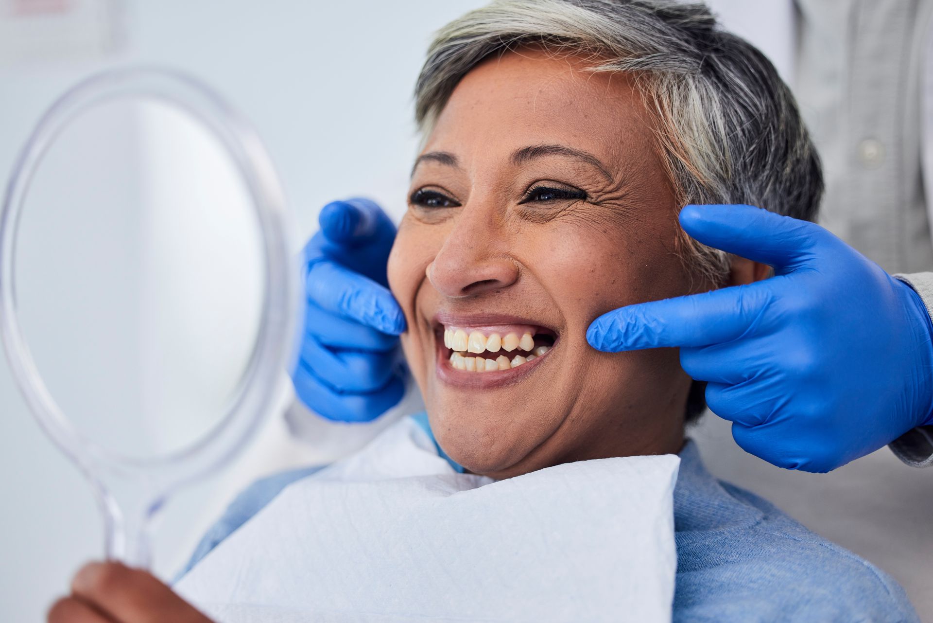 Smiling patient getting facial treatment from gloved hands in a clinic, with a handheld mirror nearby