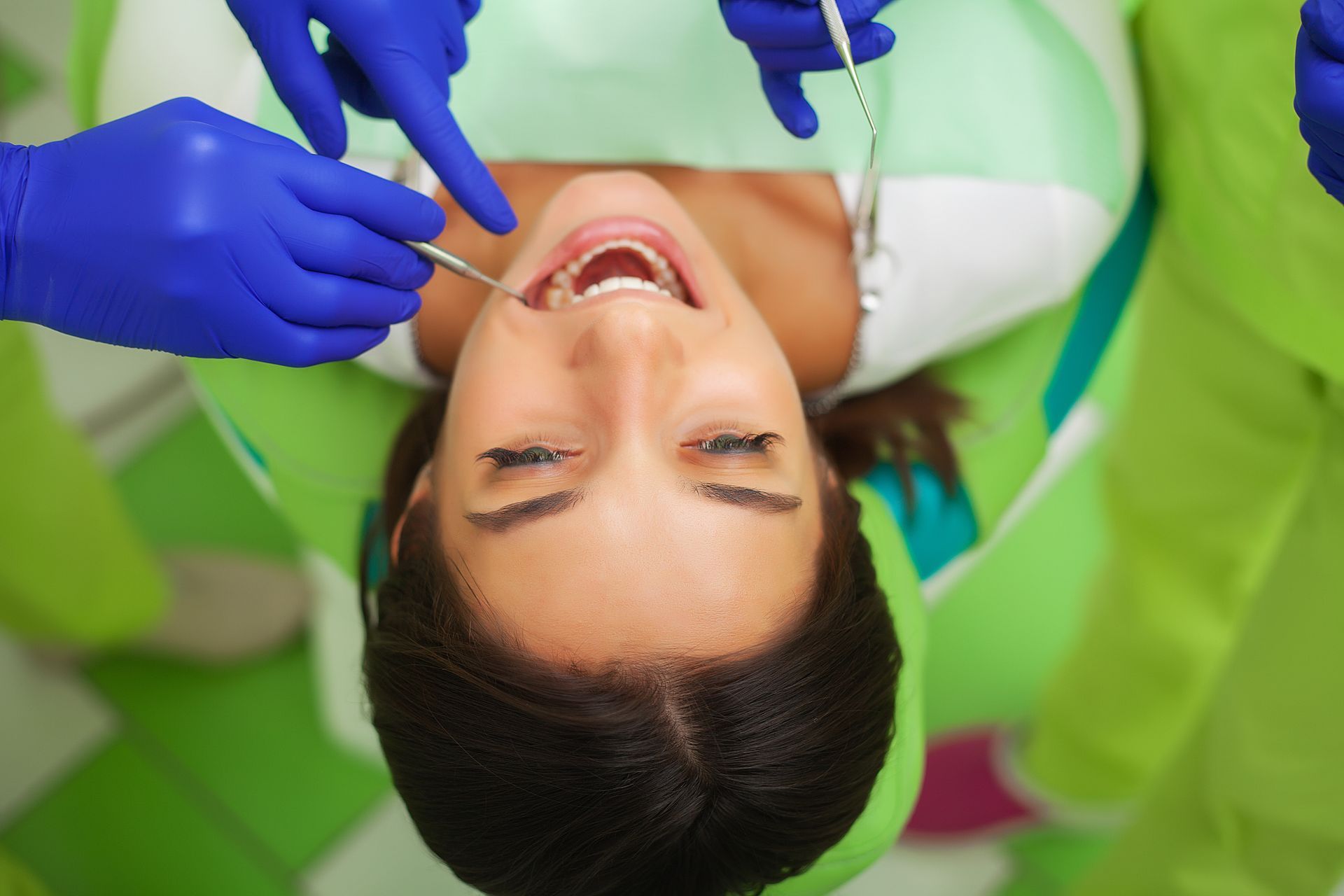Dentist examining a patient’s open mouth with blue gloves in a green dental chair