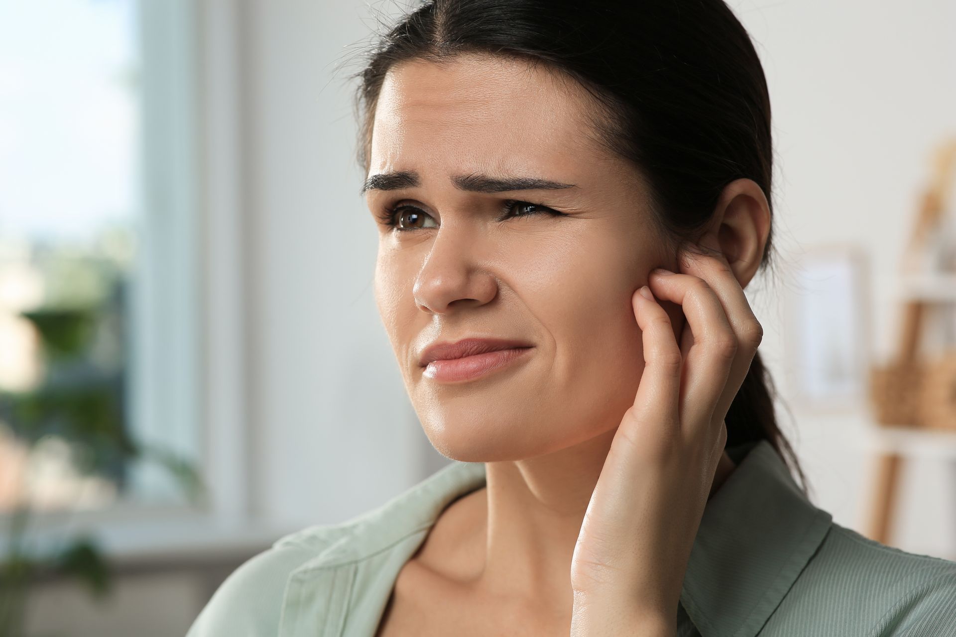 Woman touching her ear with a pained expression, indoors near a bright window.
