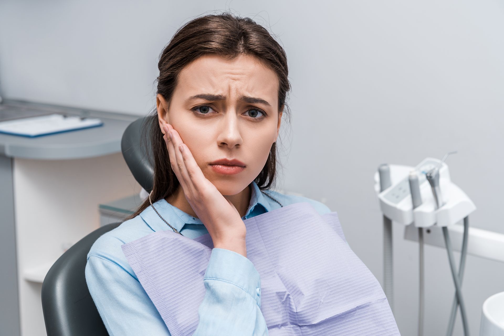 Dental patient holding her cheek in a dentist’s chair, looking worried in a clinic with dental equipment.