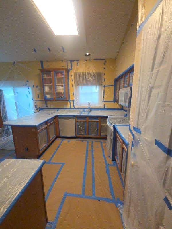 Kitchen under renovation with taped floors, covered cabinets, and a window over the sink.