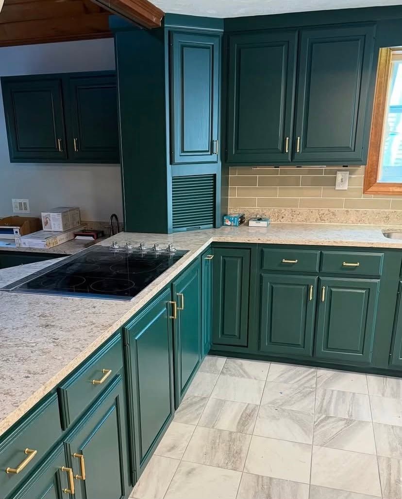 Green kitchen cabinets with gold hardware, speckled countertops, and a window over the sink.