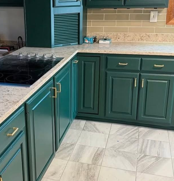 Teal kitchen cabinets with white marble countertops and black stovetop in a tiled kitchen.