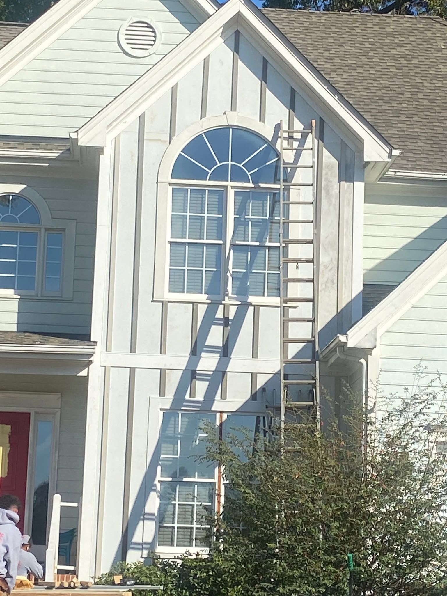 Two ladders lean against a white house, reaching a blue arched window above a bush.