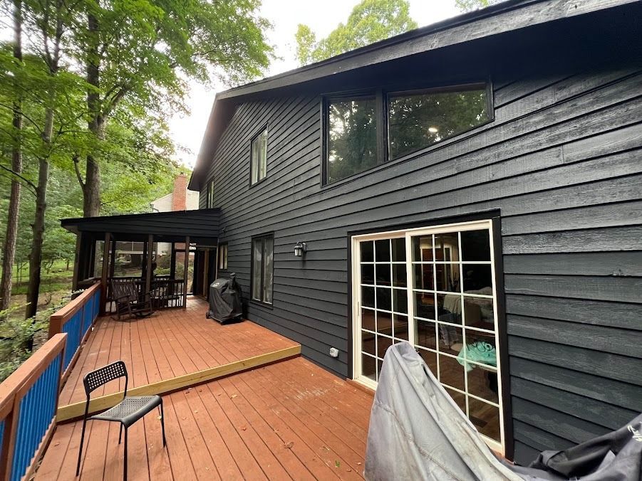Black-sided house with wooden deck, blue railing, and trees surrounding the backyard.