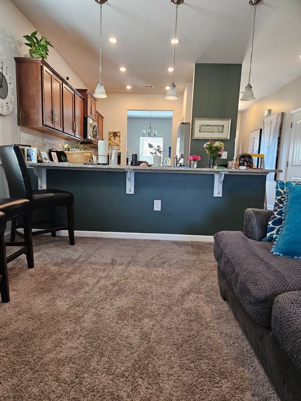 Carpeted living room with dark sectional, kitchen pass-through, wood cabinets, and pendant lights